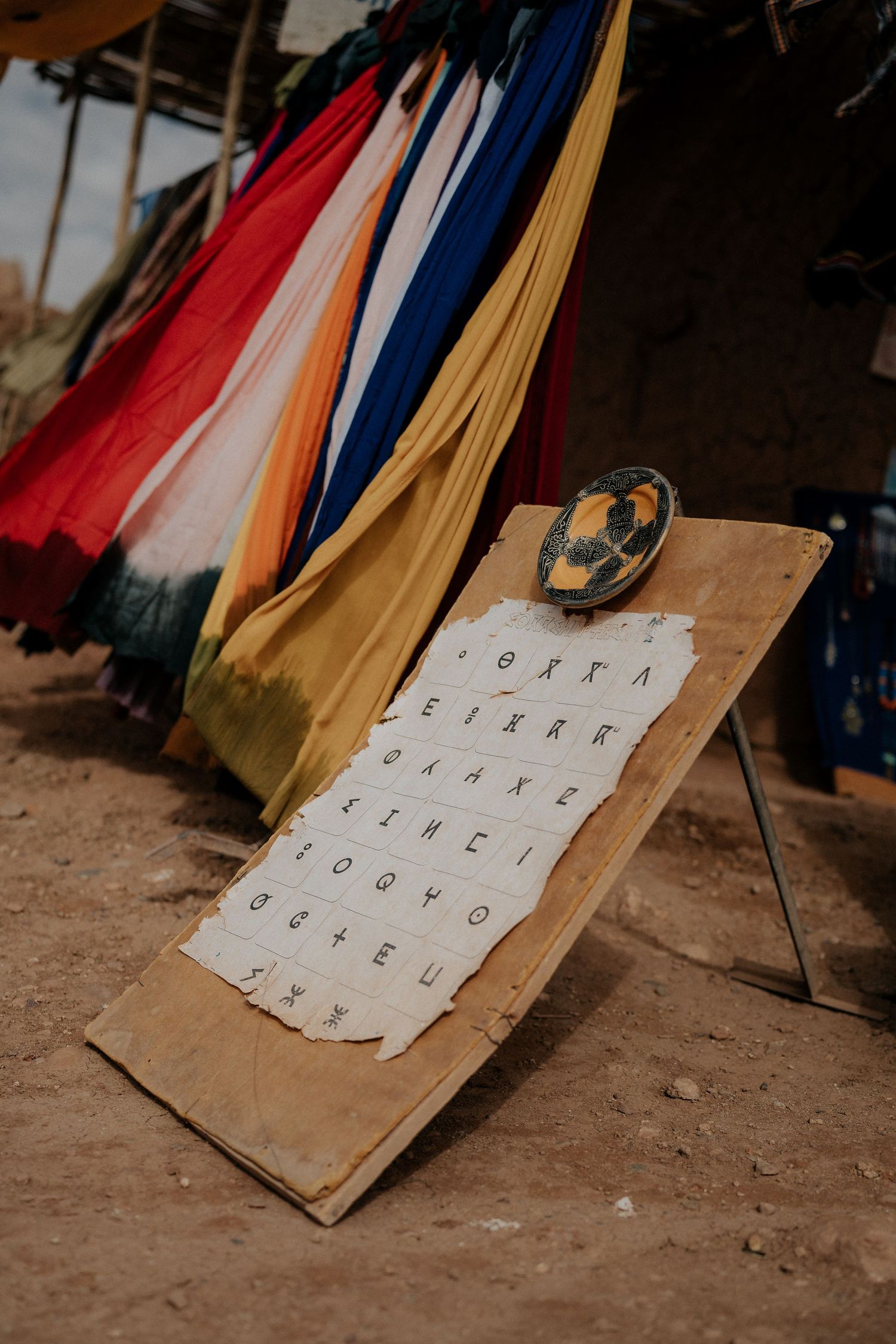 Colorful fabric hangings with alphabet chart displayed on wooden board against earthen wall.