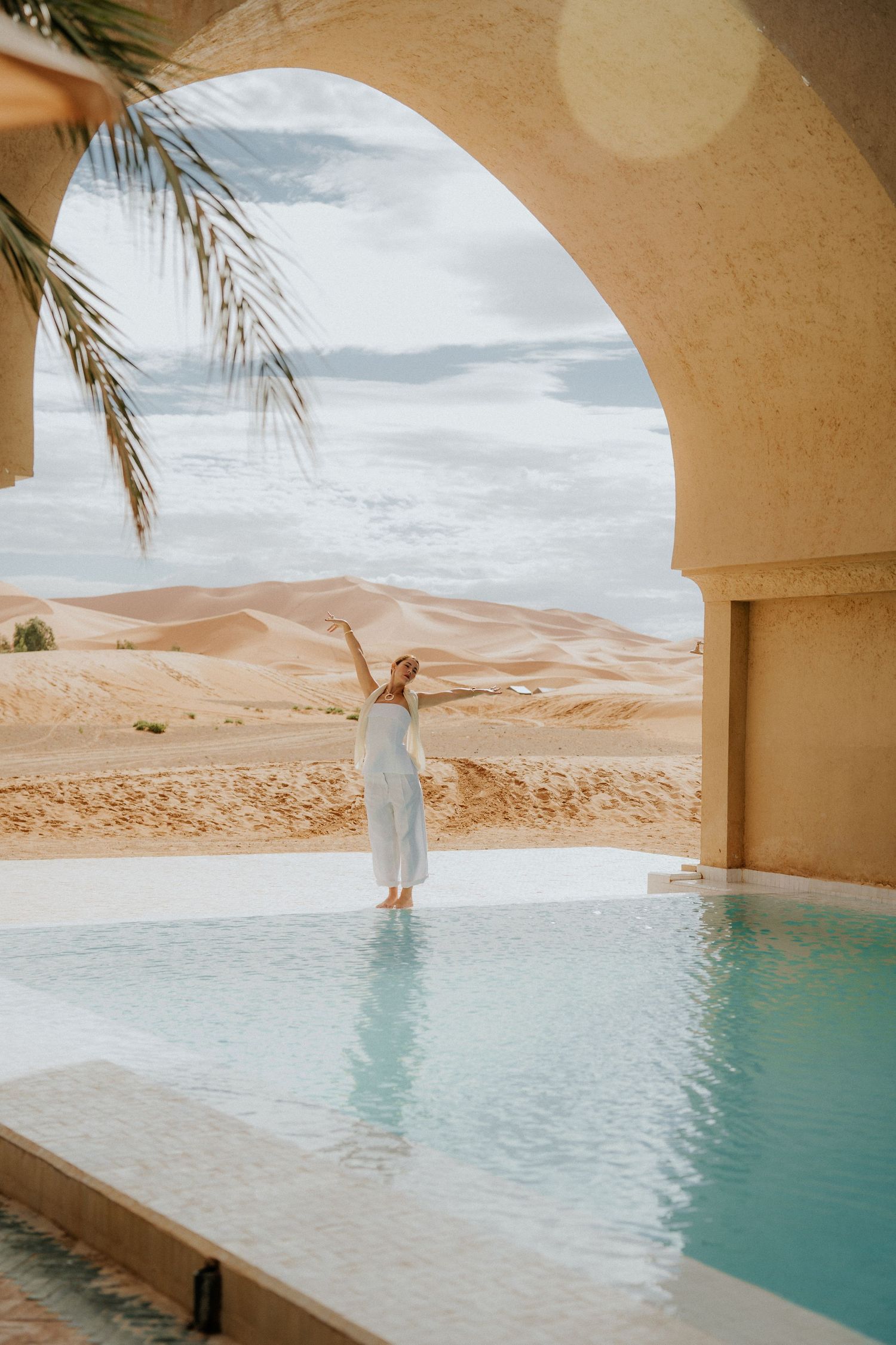 Turquoise swimming pool with arched windows frames a stunning view of desert sand dunes.