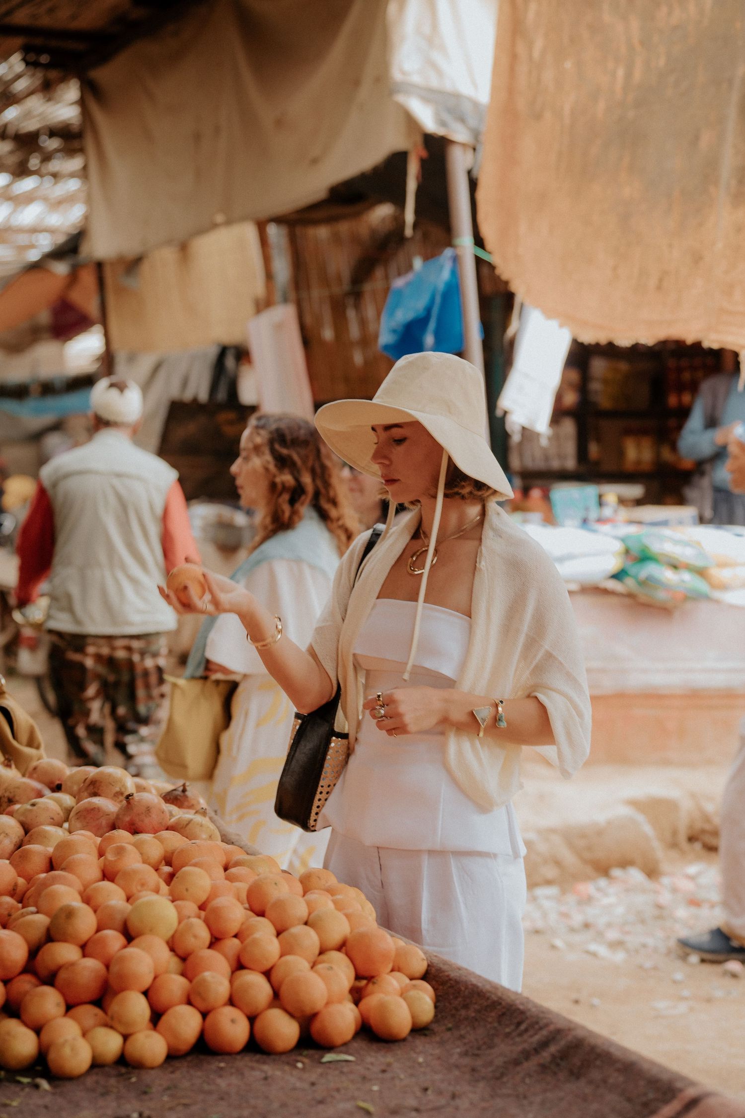 Shopper in white summer outfit browses fresh oranges at a local market stall.