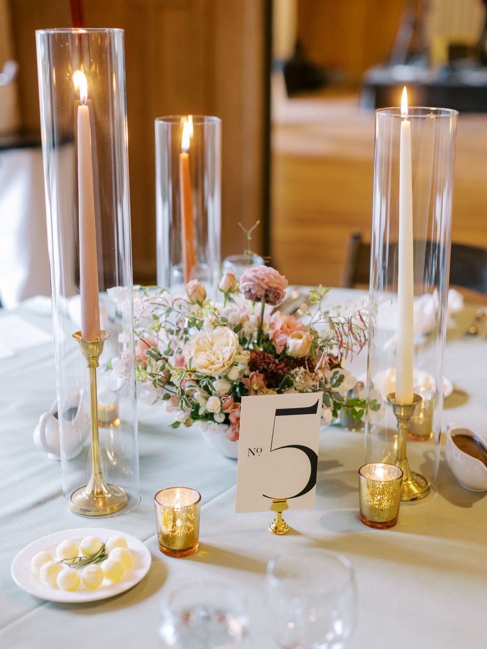 Elegant wedding table setting with tall glass candleholders, floral centerpiece, and table number 5 on white linens.