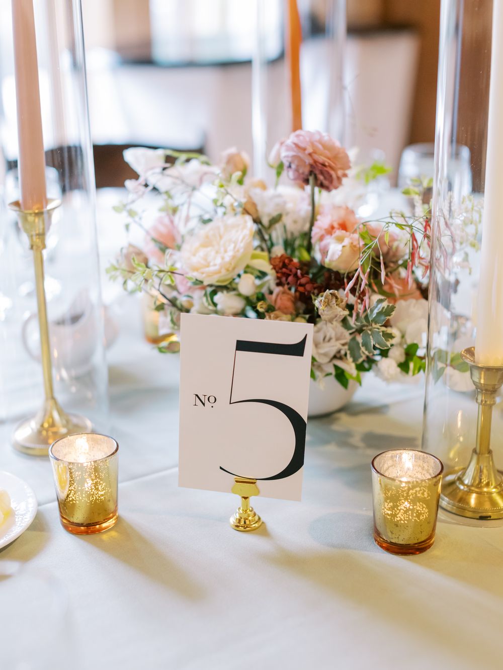 Table number 5 card displayed with delicate pink floral centerpiece and amber glass votives at an elegant reception.