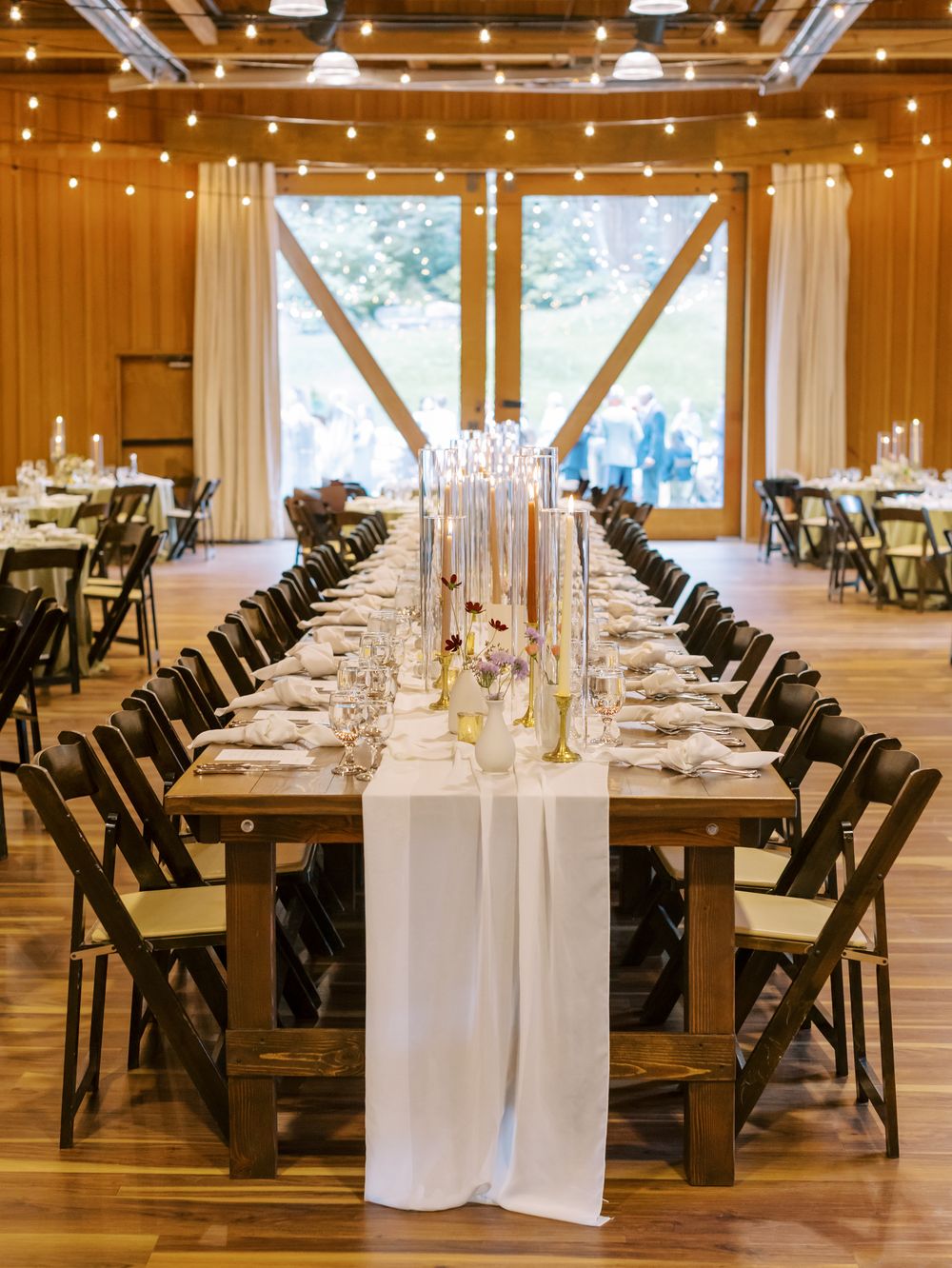 Wedding reception venue with rustic wooden beams, string lights, and long white-clothed dining tables set for an elegant event.