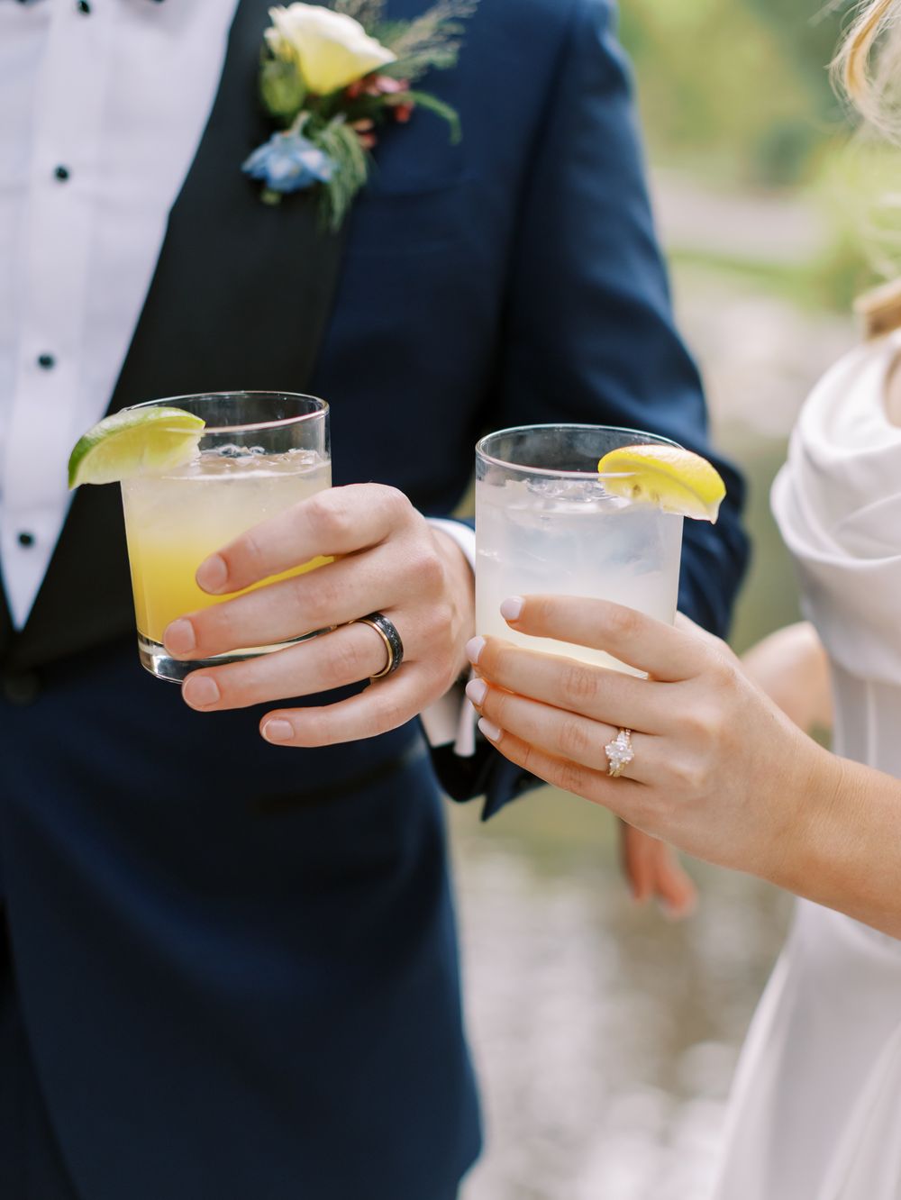 Hands holding elegant cocktail glasses with yellow drinks garnished with lemon slices at an outdoor event.