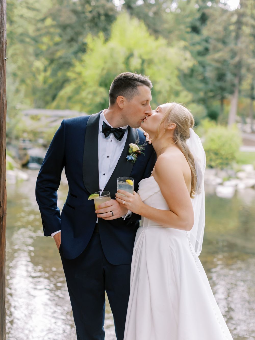 A romantic wedding couple shares an intimate moment by a lakeside in formal attire with natural scenery in the background.