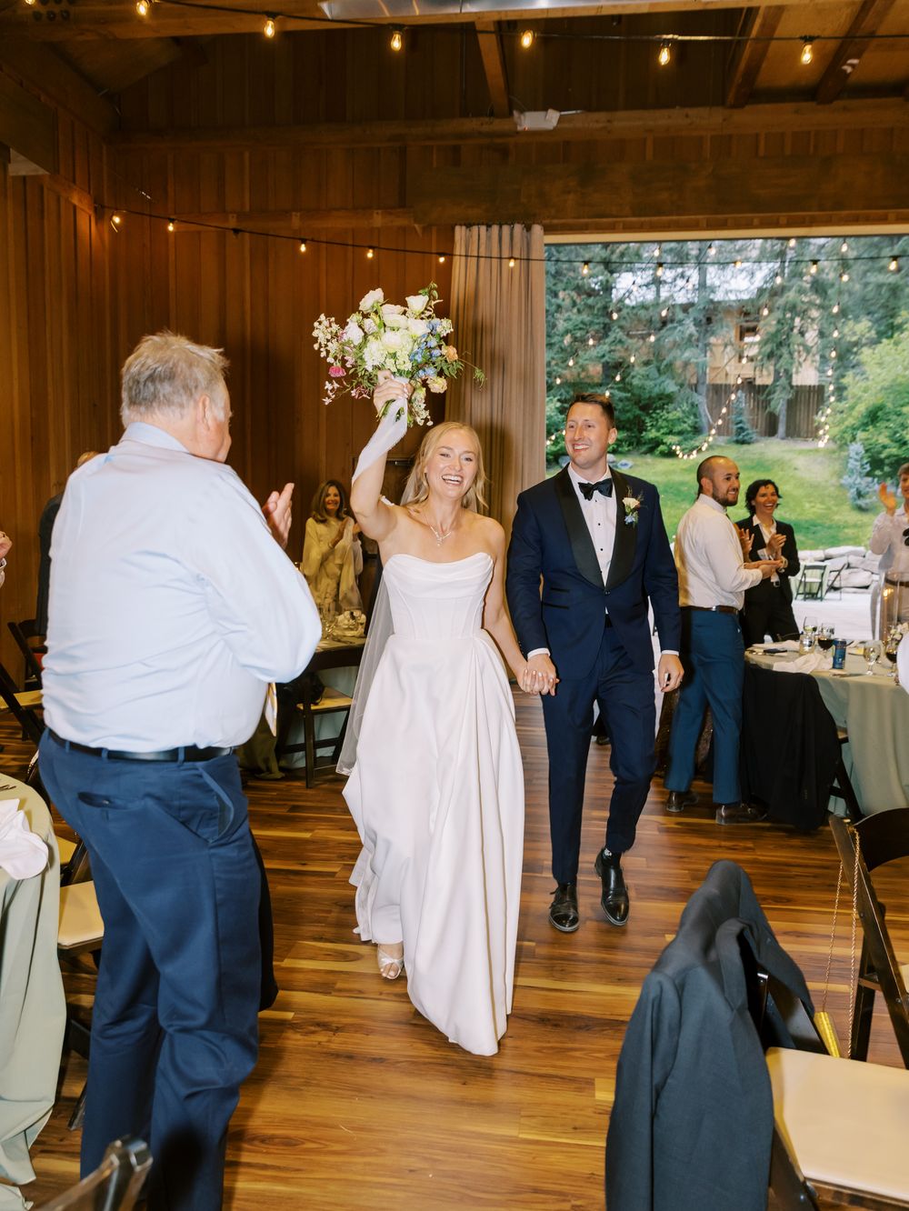 Wedding guests dancing and mingling in a rustic barn reception venue with warm lighting and wooden beams.