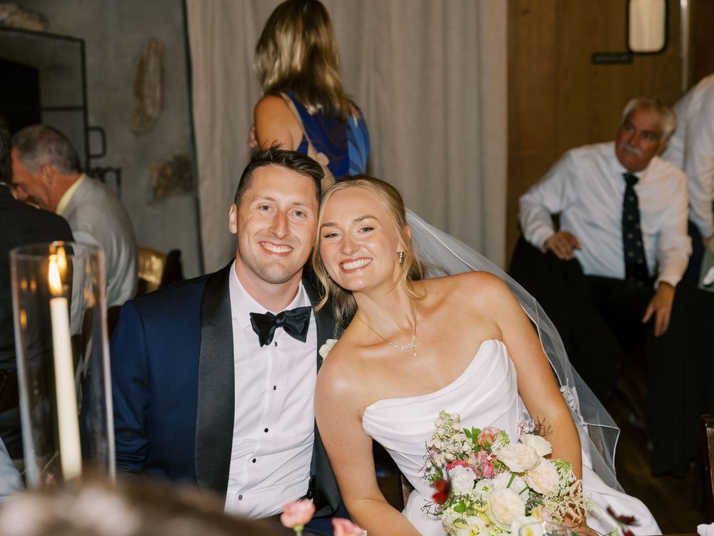 A wedding celebration with the newlyweds sitting close together at their reception, sharing bright smiles.
