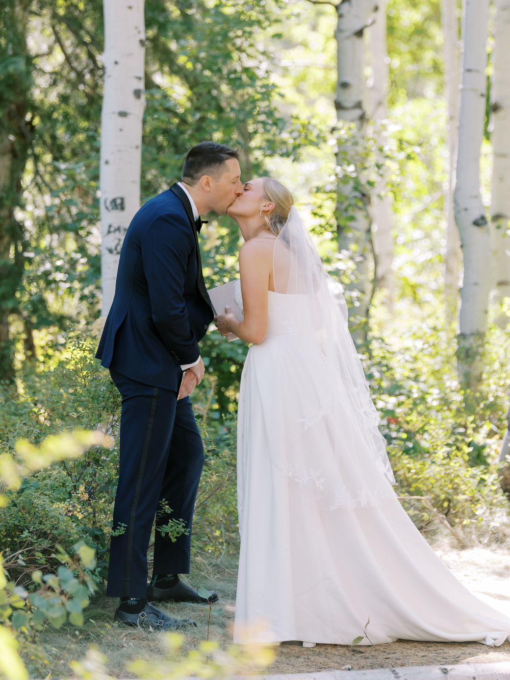 Romantic wedding moment between couple in white gown and navy suit amidst sunlit birch trees.