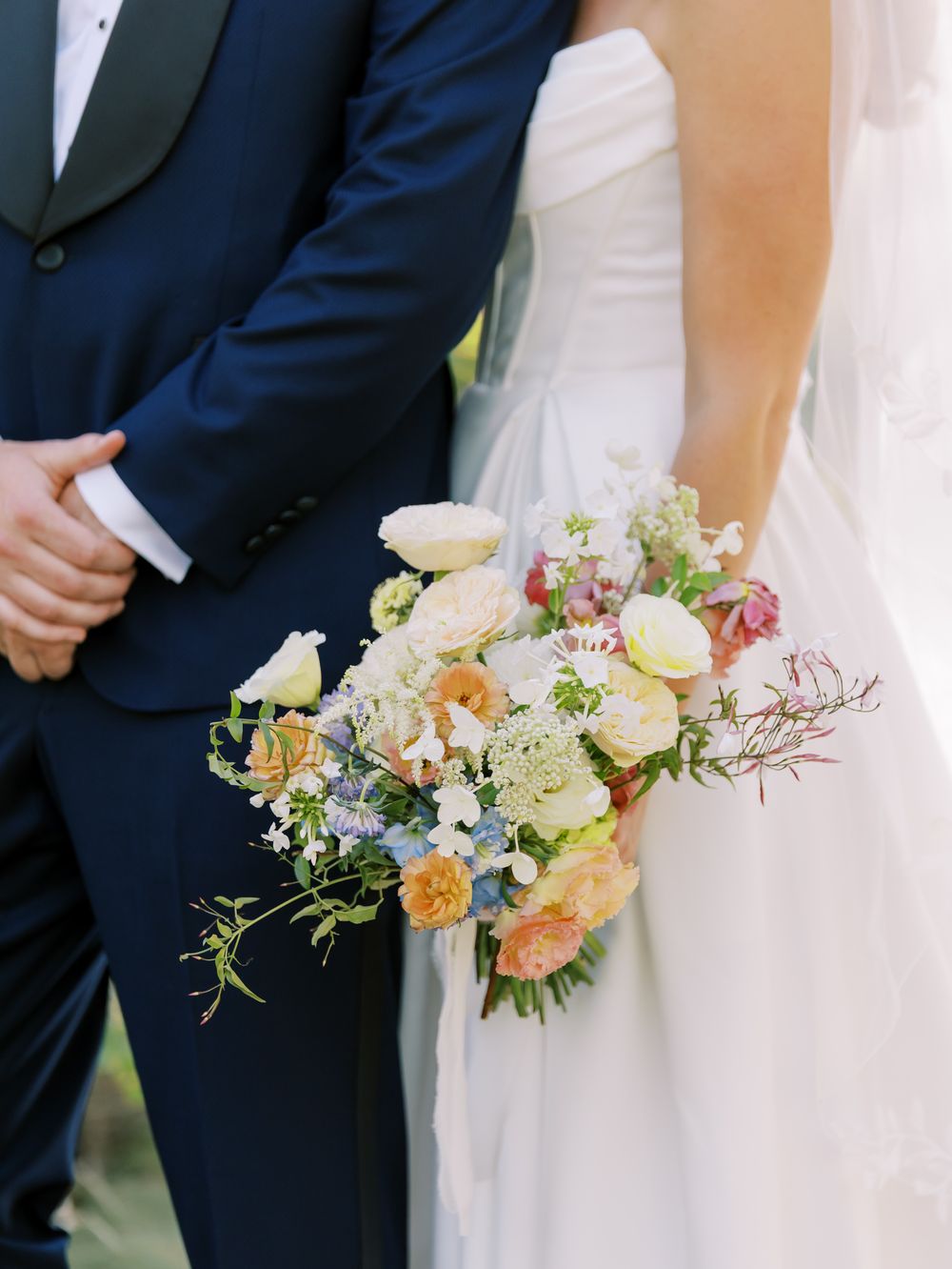 An elegant wedding bouquet of peach and cream flowers held against a navy suit and white dress.