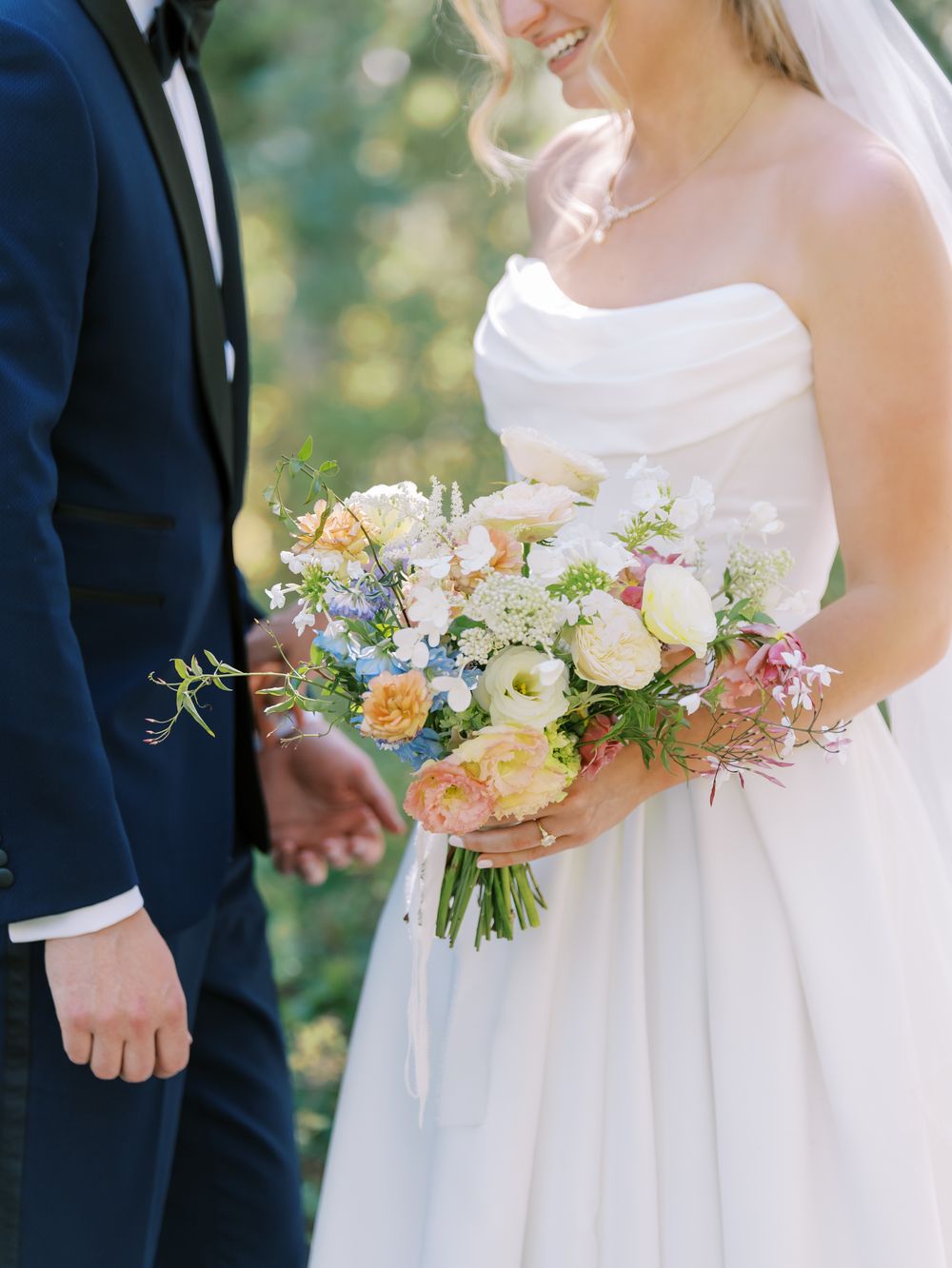 Close up of beautiful pastel wedding bouquet with peach and blue flowers held between couple in formal attire.