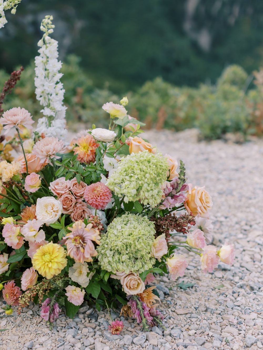 A lush garden-style floral arrangement with peach roses, hydrangeas, and delphinium on a gravel pathway.
