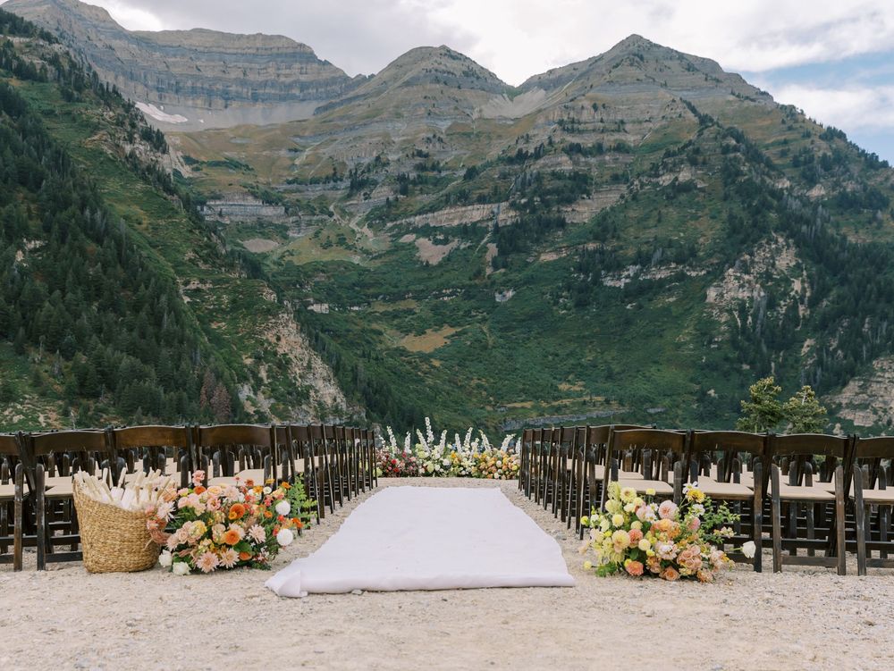 Outdoor mountain wedding ceremony setup with white aisle runner and wooden chairs framed by floral arrangements.