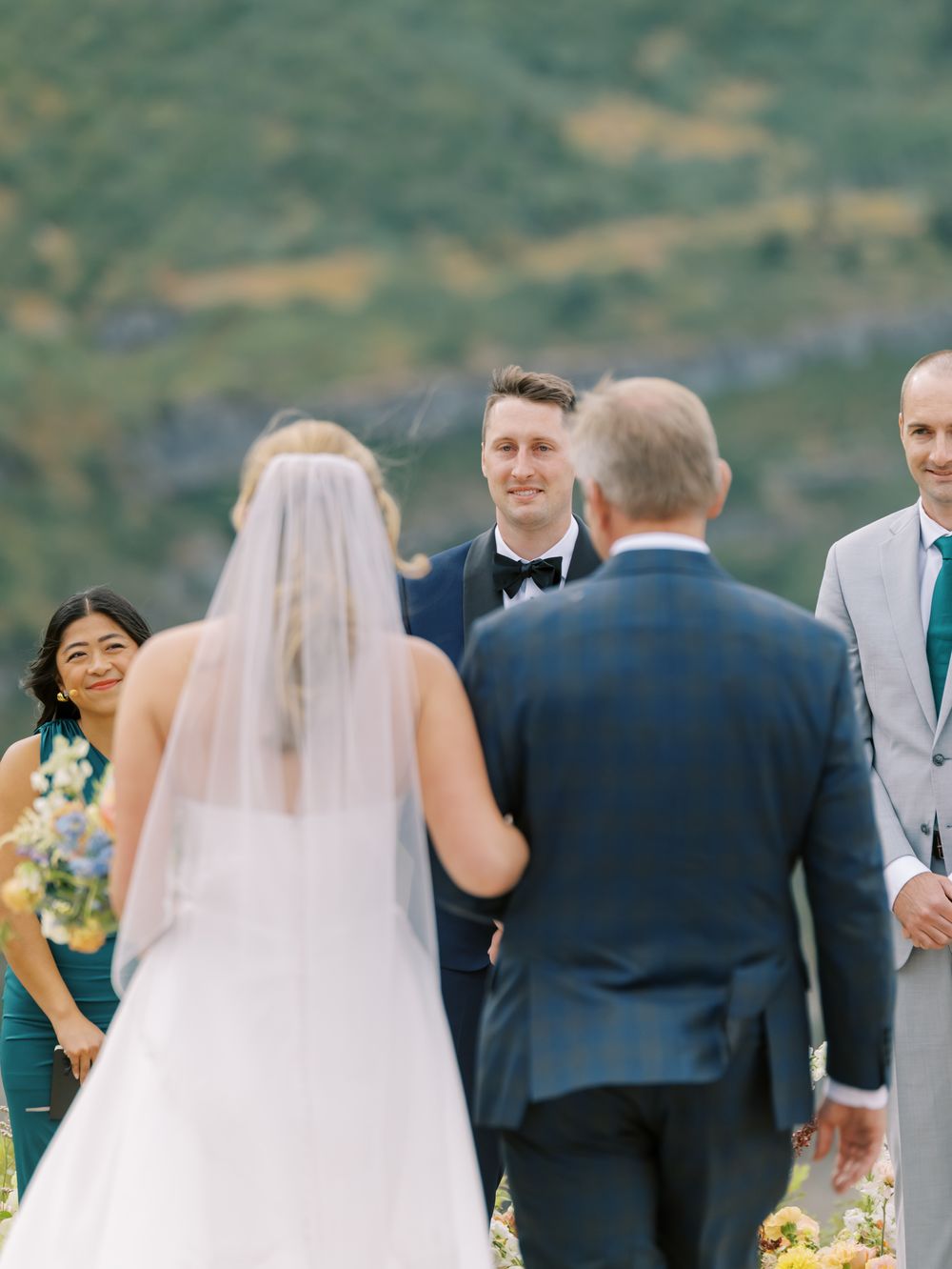 Wedding party members stand in a row outdoors during a scenic mountain ceremony with lush greenery in background.