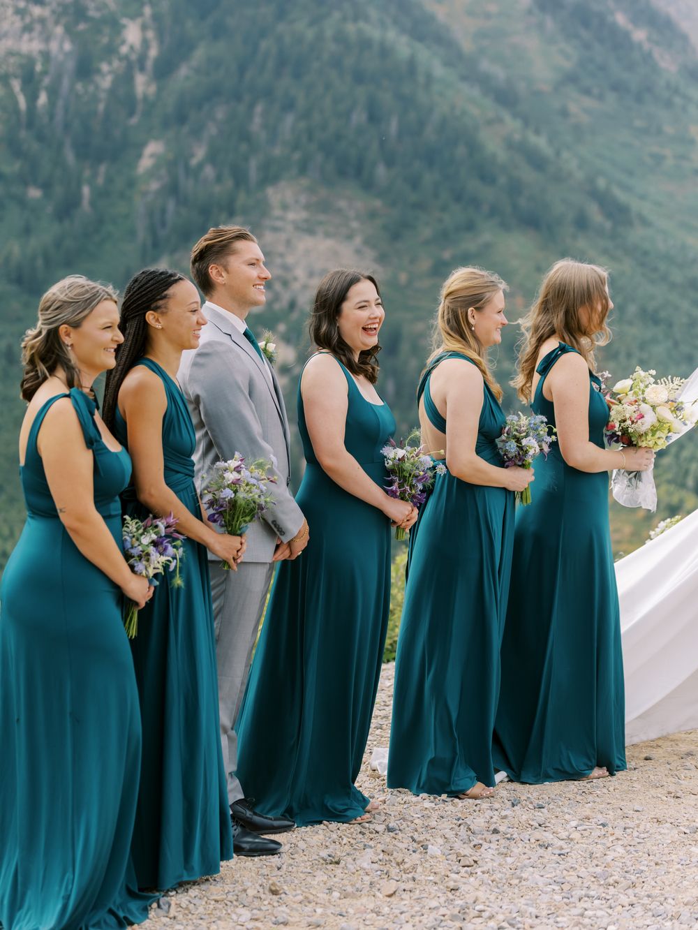 Wedding party in teal bridesmaid dresses lined up against a scenic mountain backdrop.