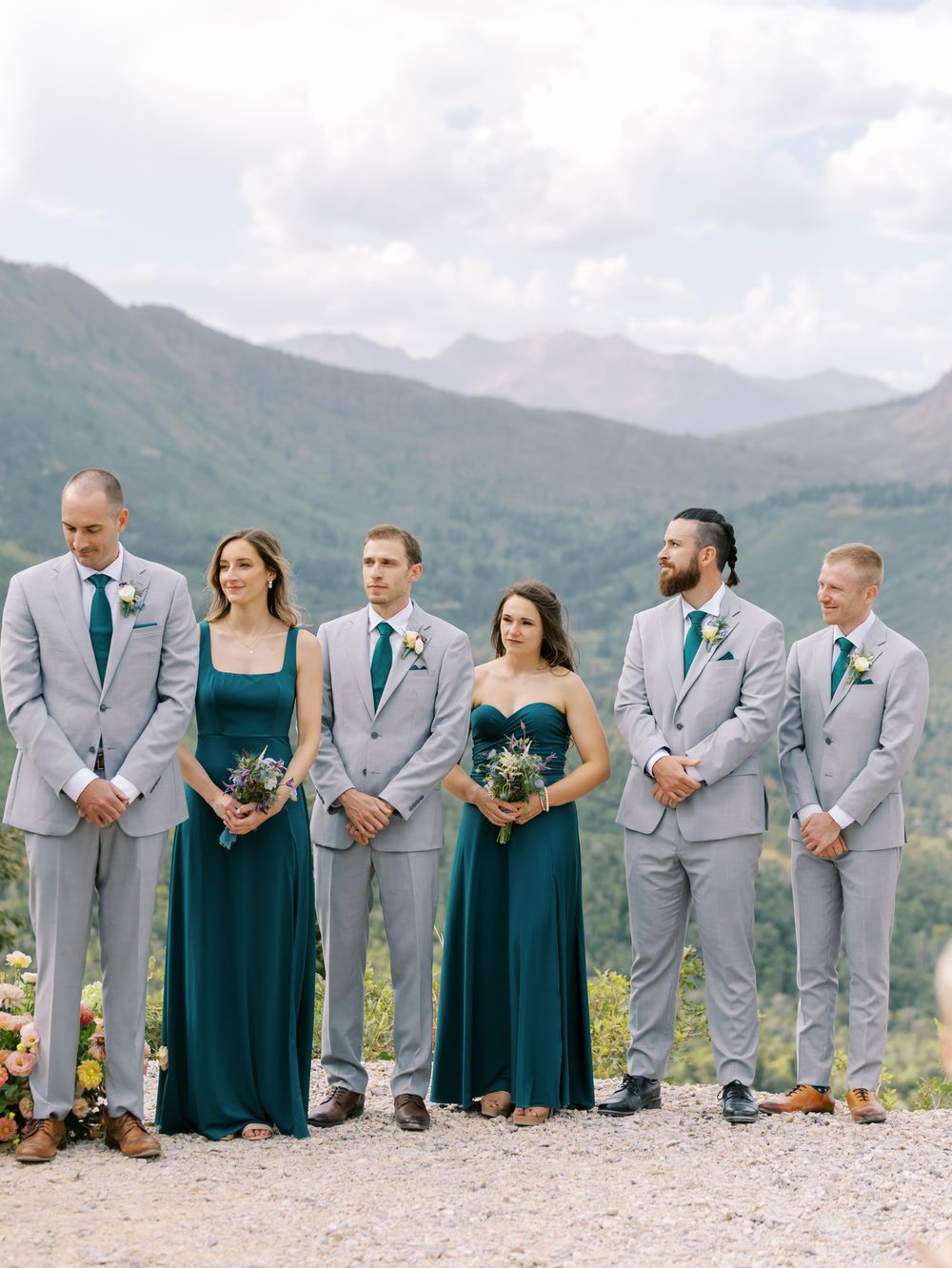 Wedding party in gray suits and teal dresses pose against a mountain landscape backdrop.
