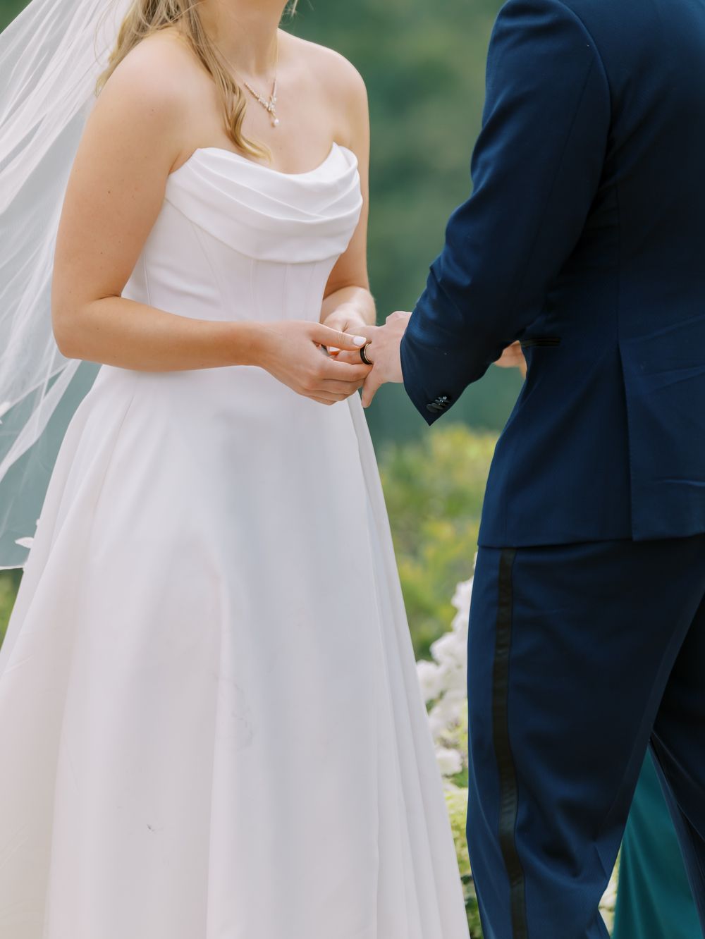 Close-up of bride and groom holding hands during wedding ceremony with white dress and navy suit visible.