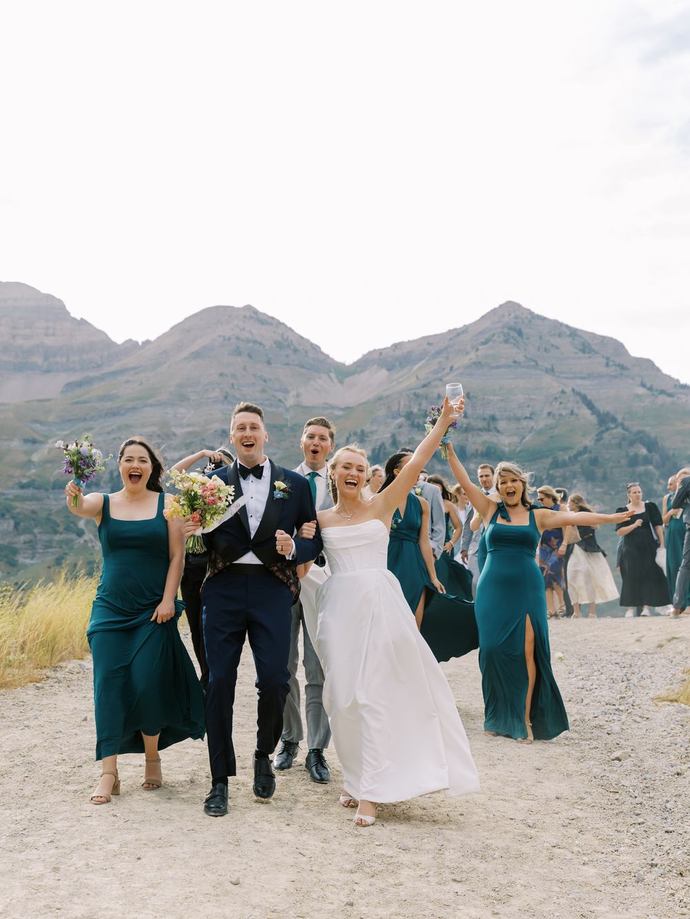 Wedding party celebrating with arms raised against dramatic mountain scenery.