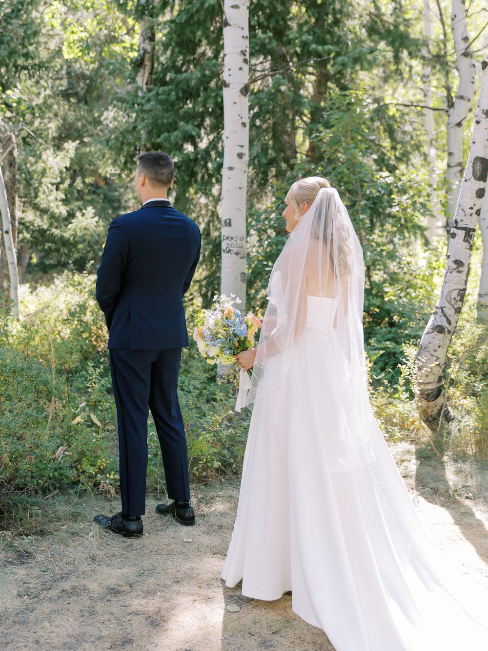 A wedding couple stands among birch trees, with the bride in a flowing white gown and veil.