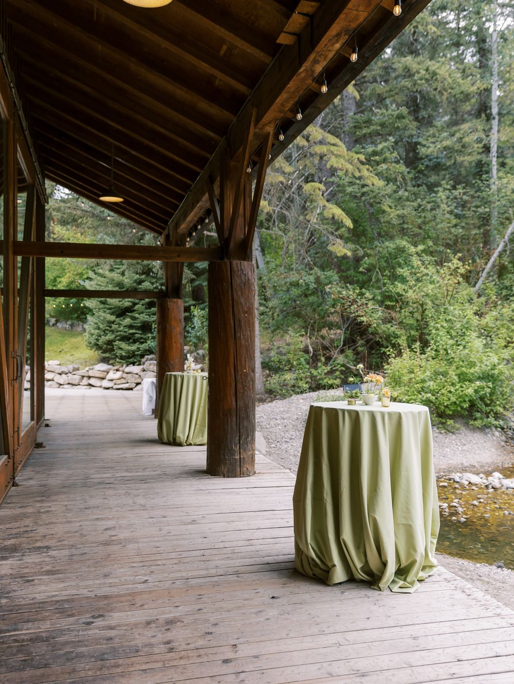 Rustic wooden covered porch with lime green tablecloths on round tables set against a forested backdrop.