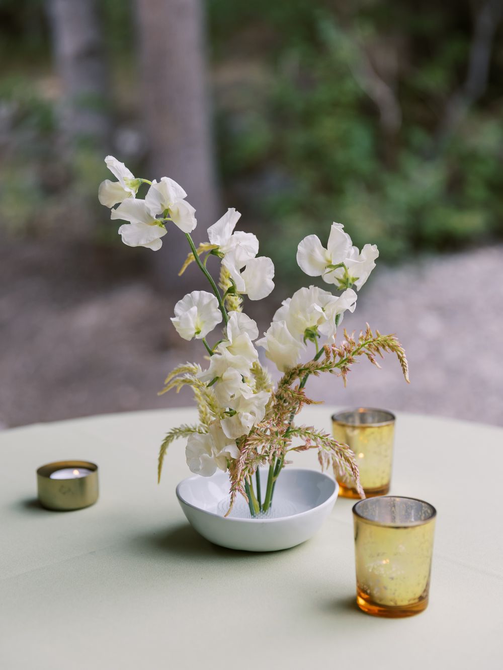 White orchid flowers arranged in a small bowl with delicate ferns and amber votives on a pale table setting.