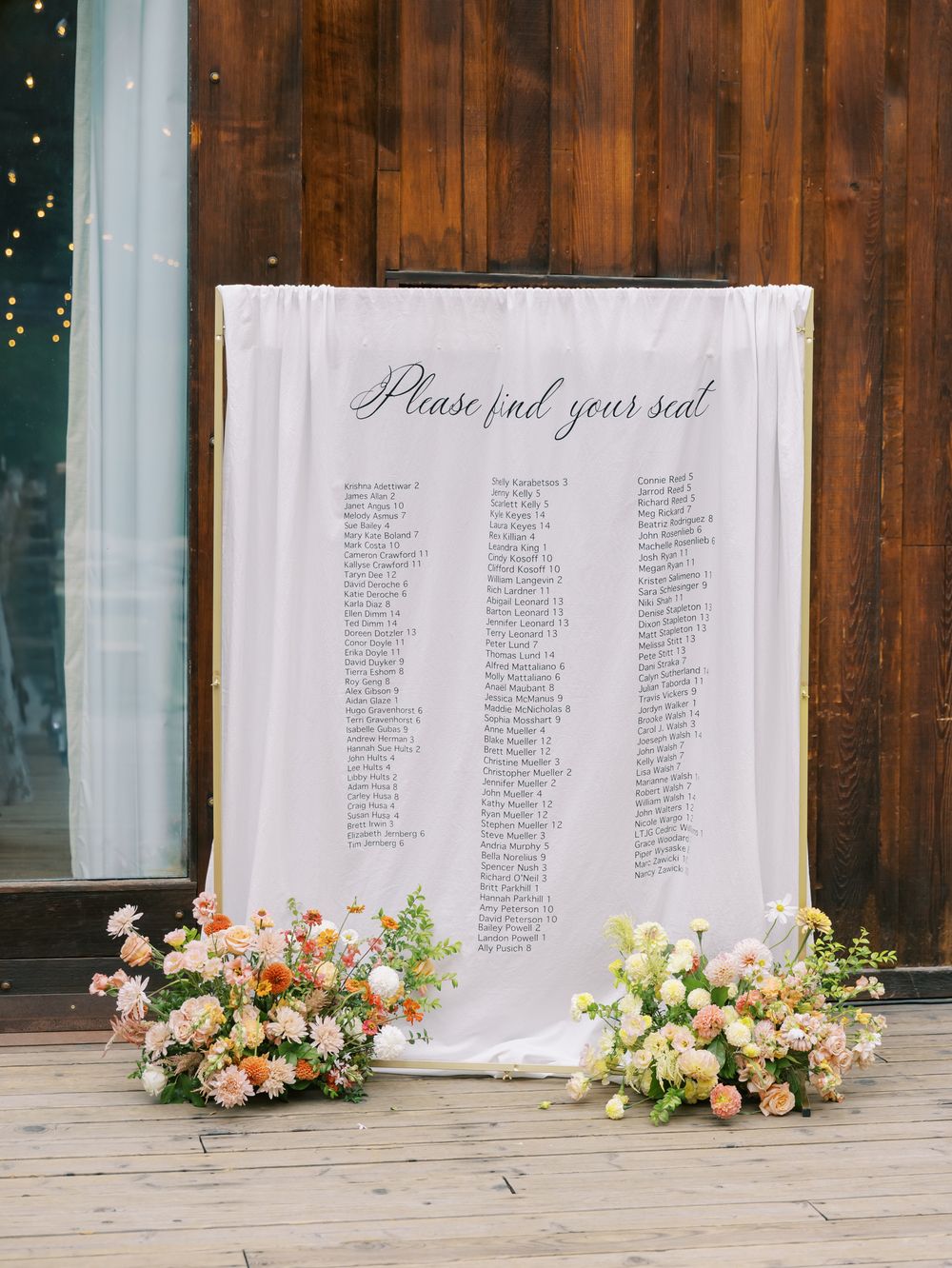 An elegant wedding seating chart display with flowing white fabric backdrop and coral floral arrangements on a wooden floor.