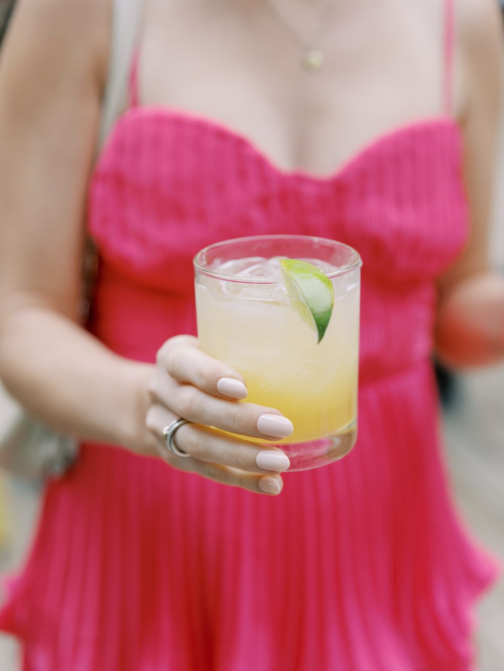 Person in bright pink dress holds a refreshing cocktail garnished with cucumber in a rocks glass.