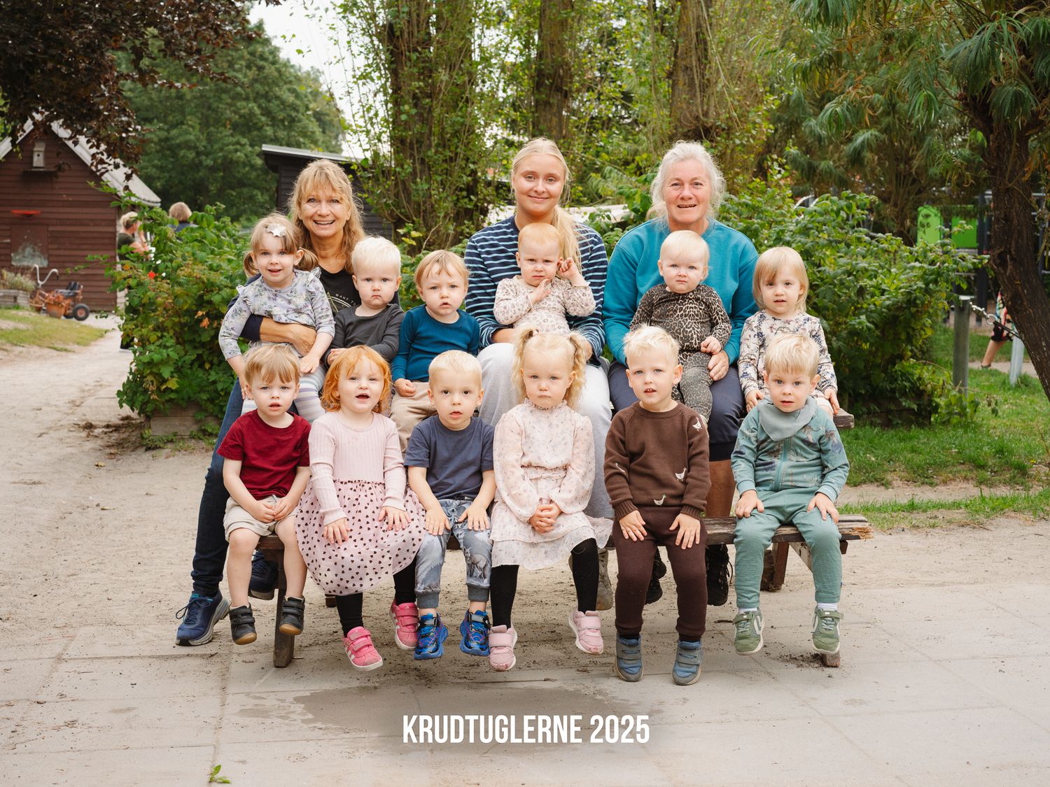 Group of children crouching in a line on a dirt path outdoors with trees and greenery in the background.