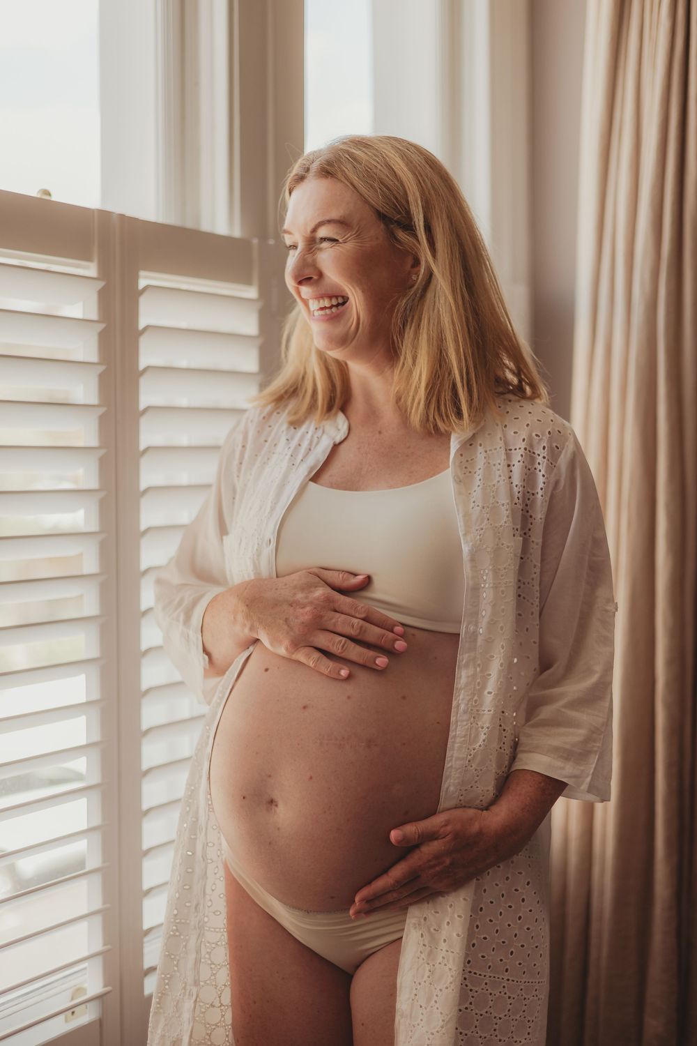 Expectant mother in white bodysuit and cardigan laughs joyfully while standing by window with natural light.