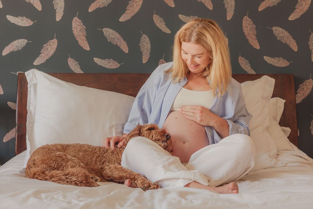Pregnant woman relaxes on bed with brown dog while wearing blue button-up shirt.