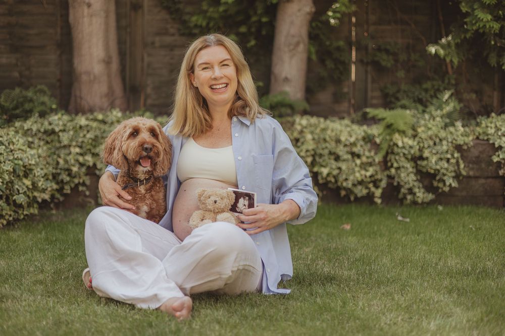 Maternity photo in garden setting with small brown dog sitting beside pregnant woman.