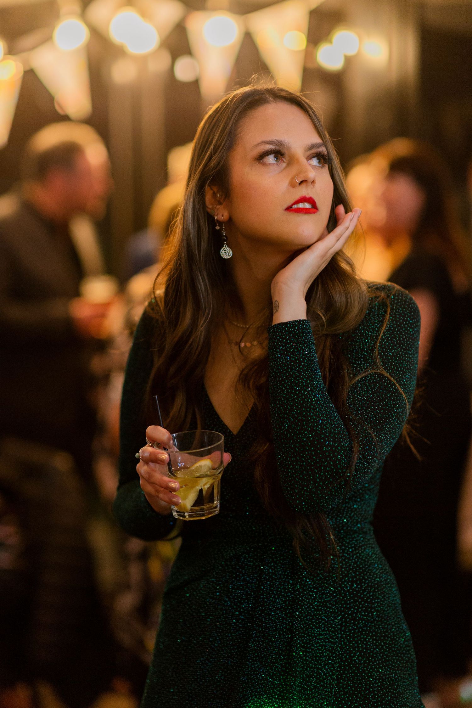 A person in a green sweater holds a cocktail glass while sitting thoughtfully at a dimly lit bar.