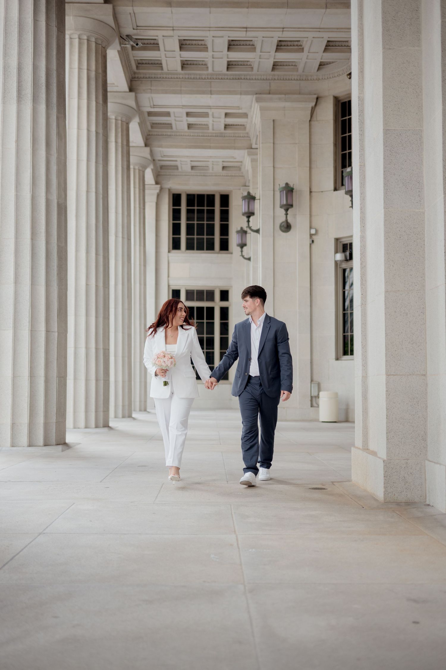 An elegantly dressed couple walks hand in hand through a classic colonnade with coffered ceilings and stone pillars.
