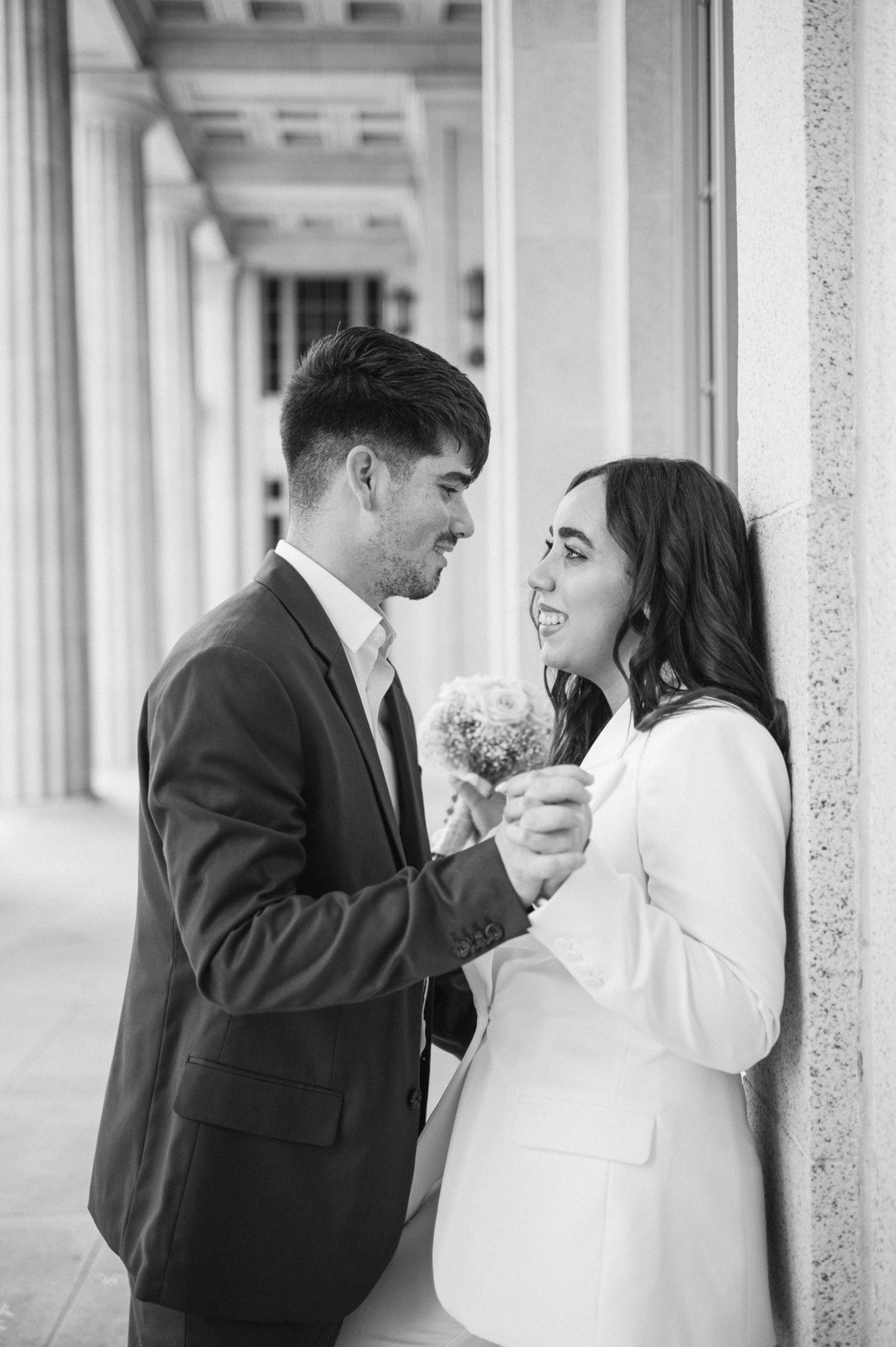A romantic black and white portrait of a couple sharing a joyful moment in an elegant columned hallway.