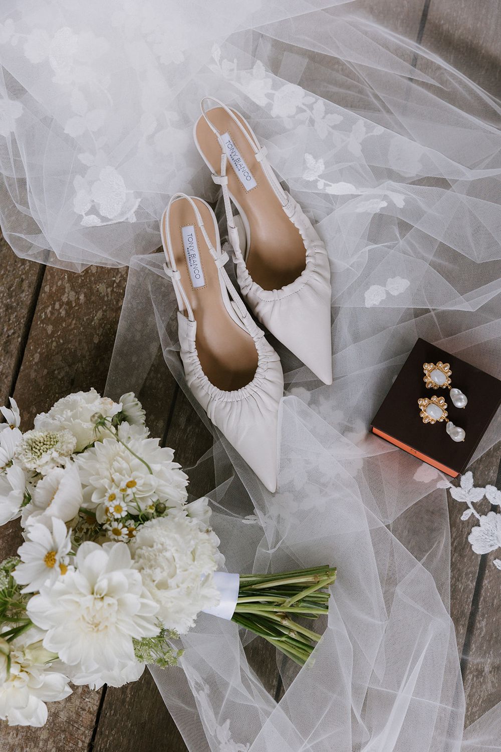 A delicate white wedding veil lies beside elegant white flowers and ivory wedding shoes on a rustic stone surface.