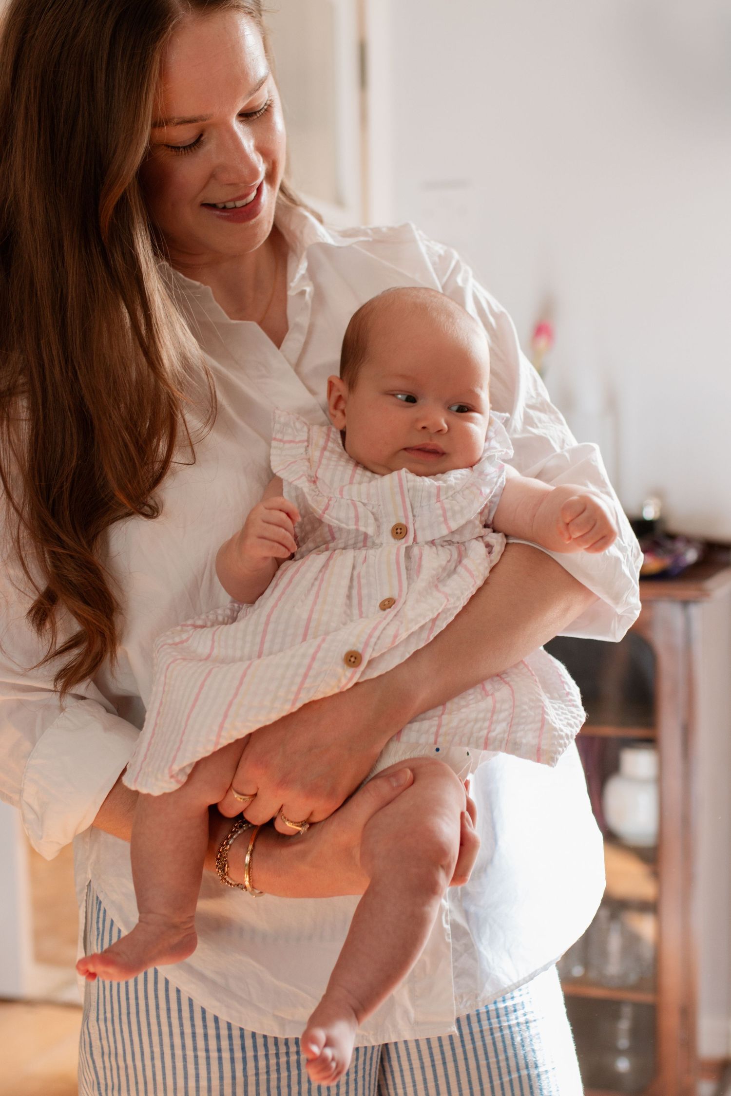 Parent holding infant wearing white clothing while standing in sunlit room.