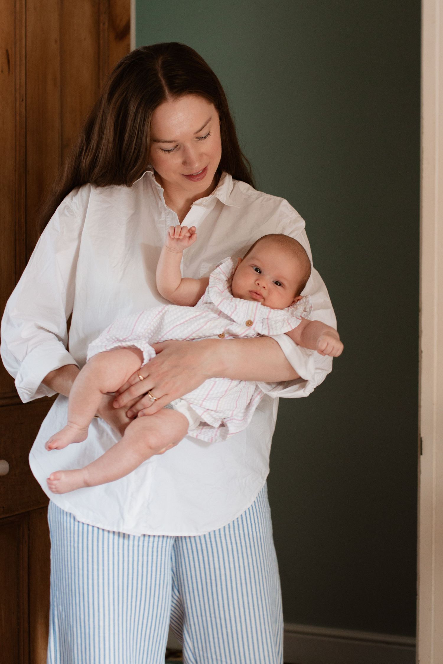 Parent holding baby against dark green wall wearing striped pants and white shirt.