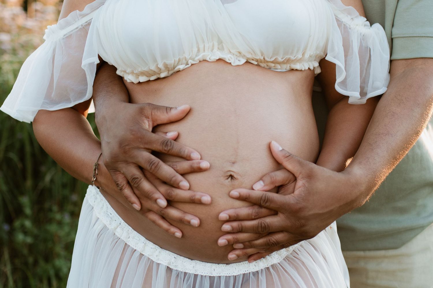 Close-up photos of hands cradling pregnant belly in white dress.