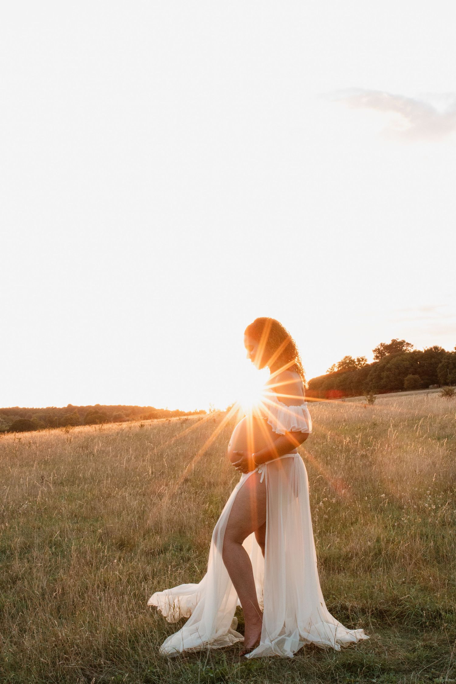 Ethereal maternity portrait with sunburst through flowing white dress in meadow.