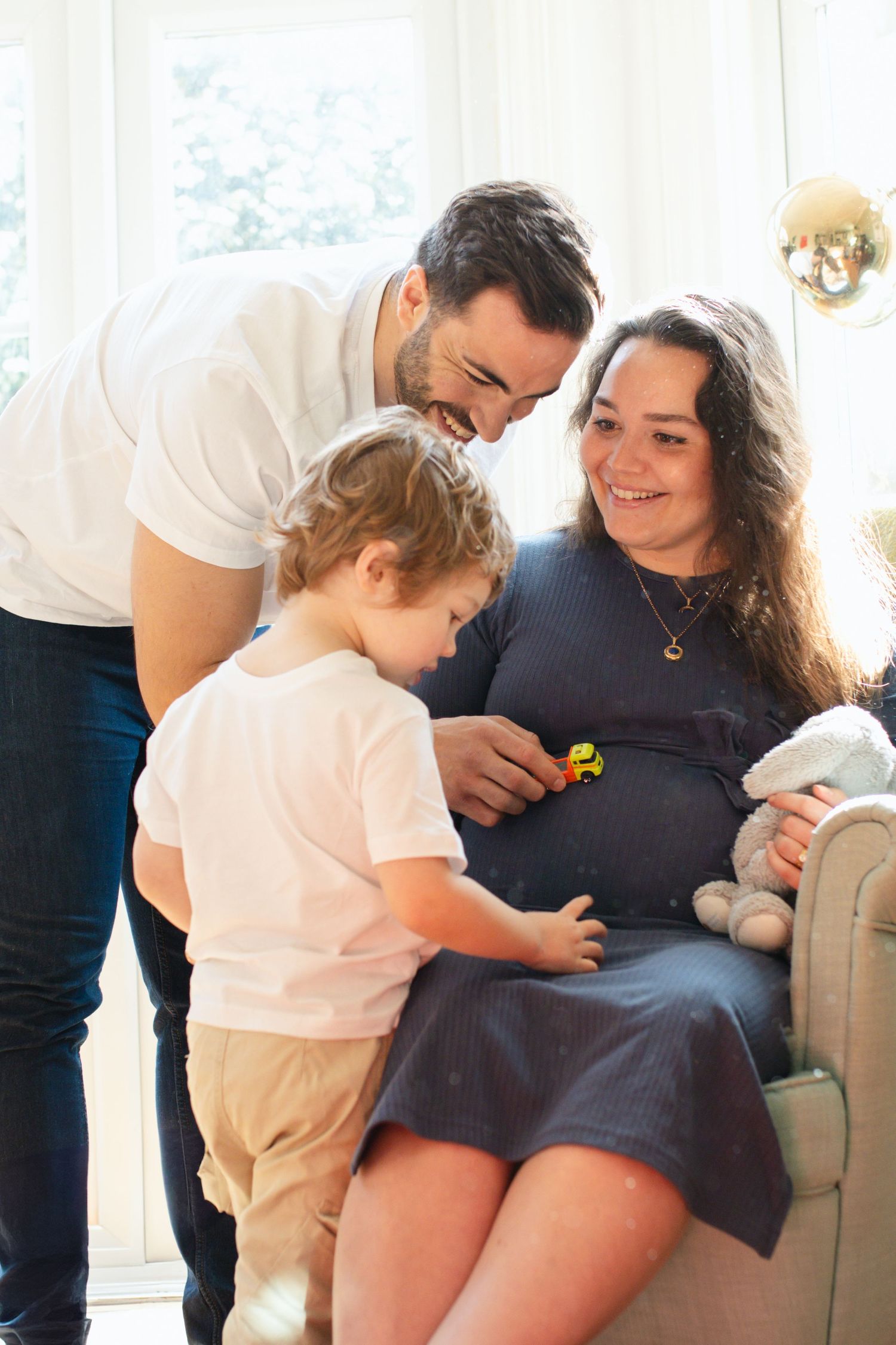 Family moment with toddler touching pregnant belly while sitting on couch.