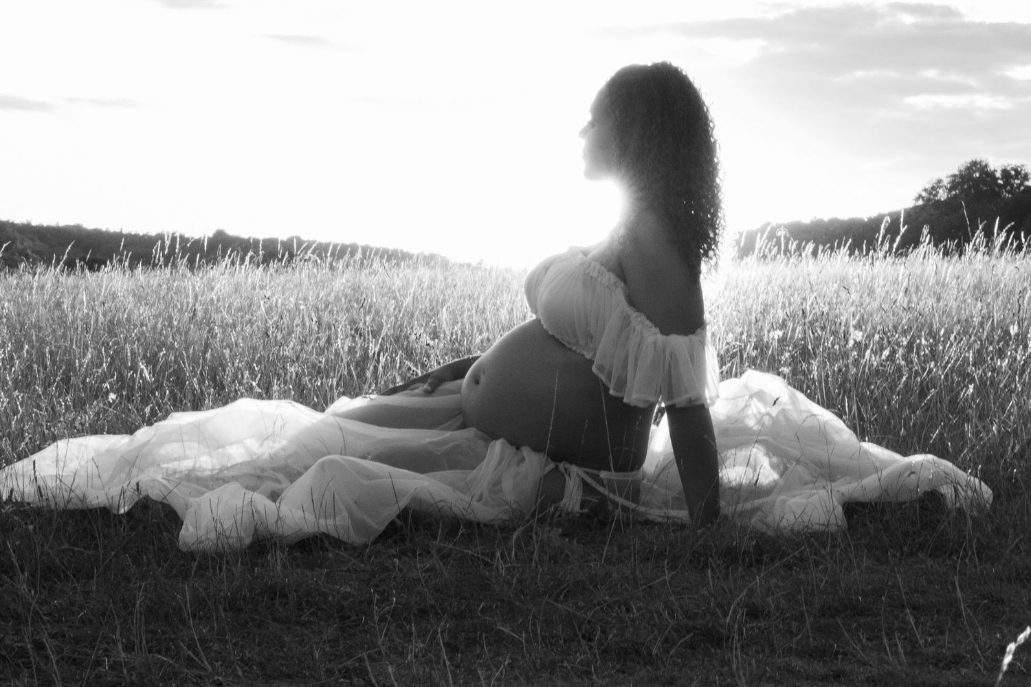 Black and white maternity photo of woman sitting in field at sunset.