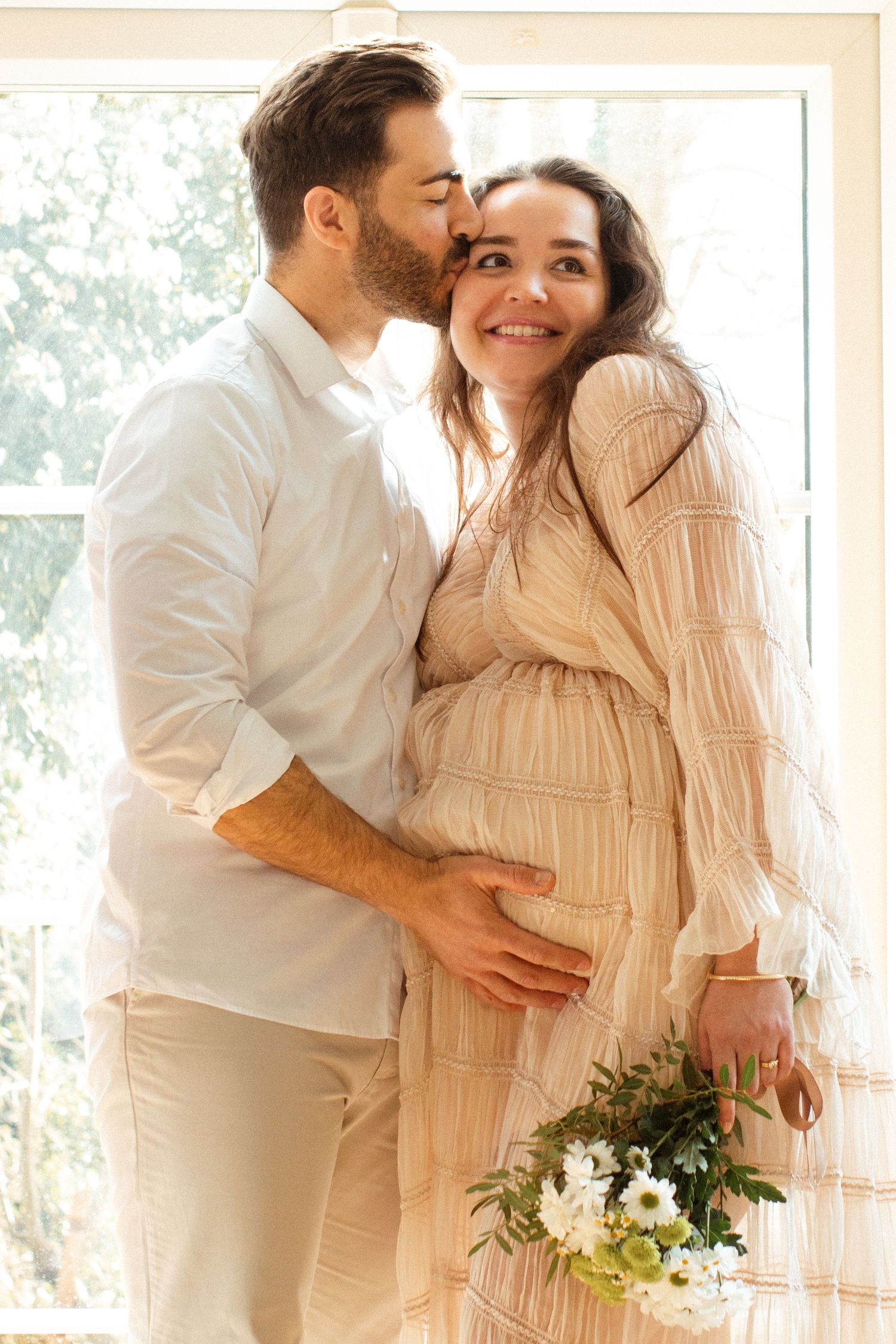 Intimate maternity portrait with couple embracing by window.