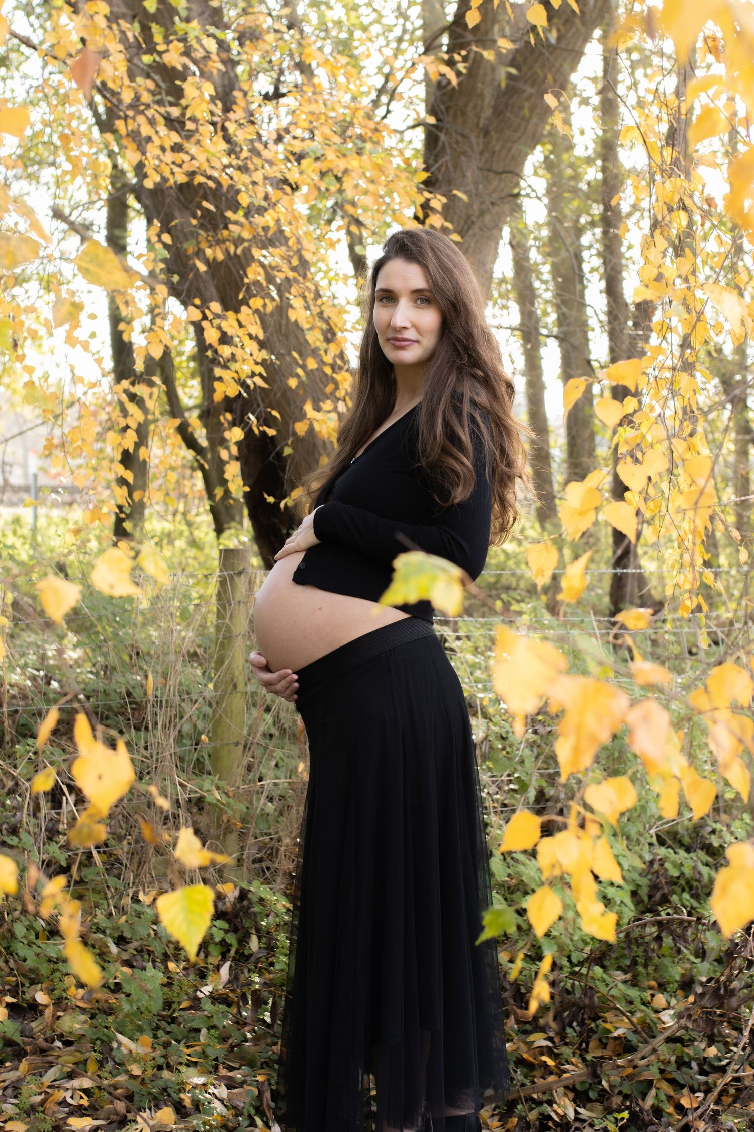 A figure stands among fall foliage wearing a flowing black dress in dappled sunlight.