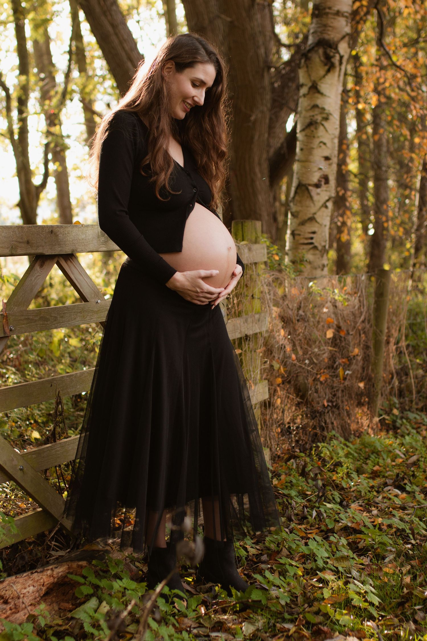 A person in a black dress poses against a rustic wooden fence in a wooded autumn setting.