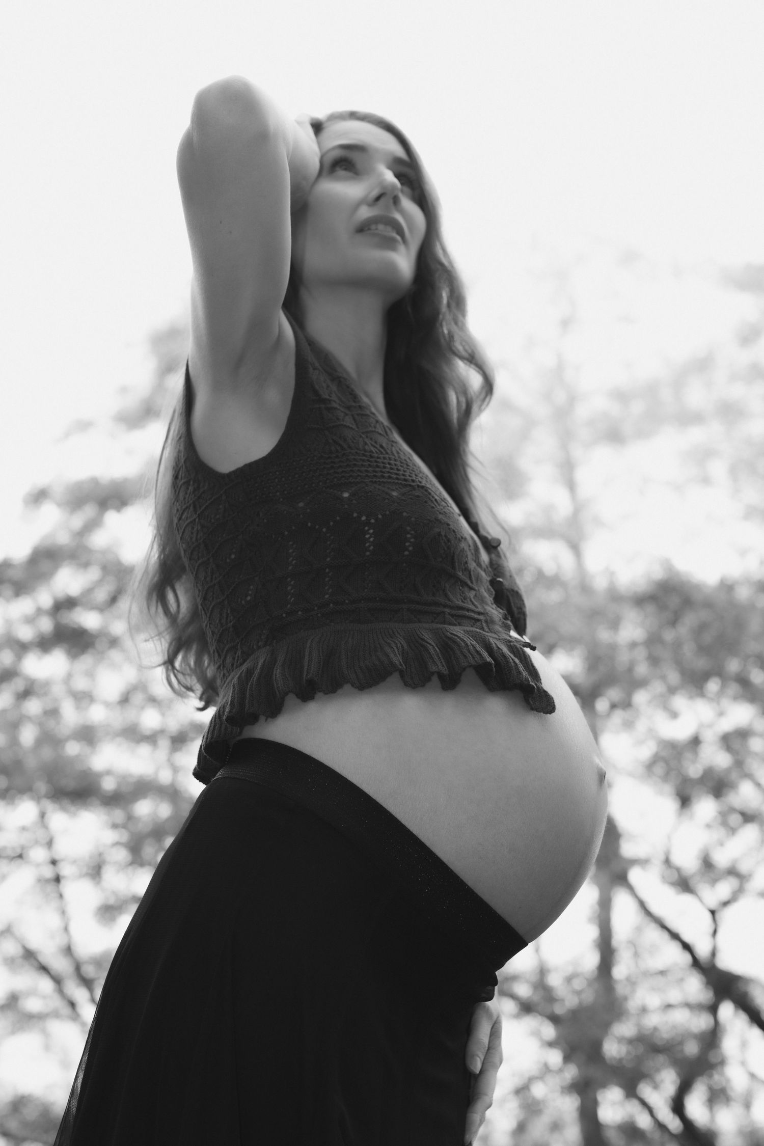 Artistic black and white upward angle portrait of a pregnant woman against a bright sky.