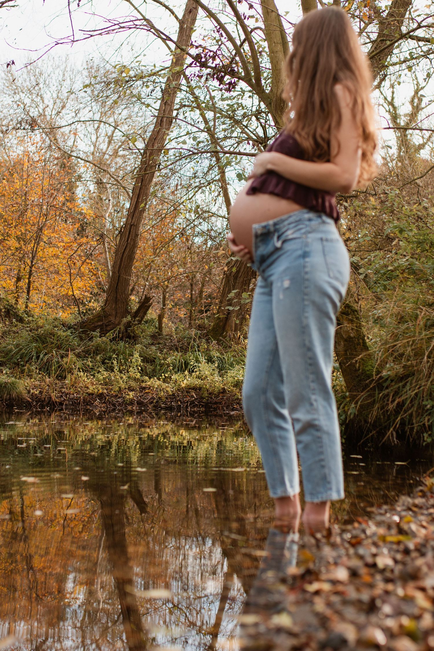 Person in denim stands by a reflective pond surrounded by autumn foliage.