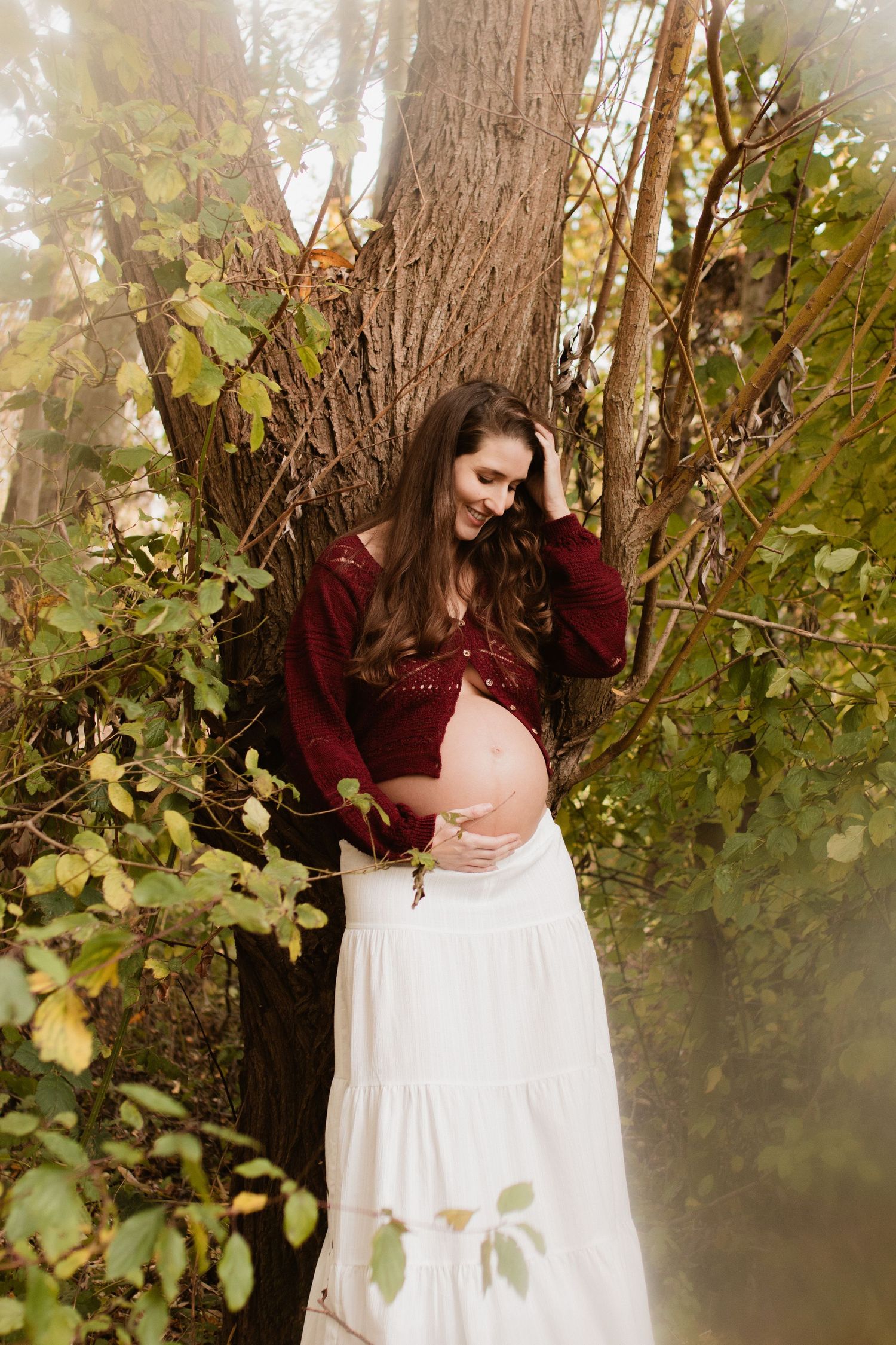 Ethereal maternity portrait in misty forest with woman in burgundy top and white skirt.