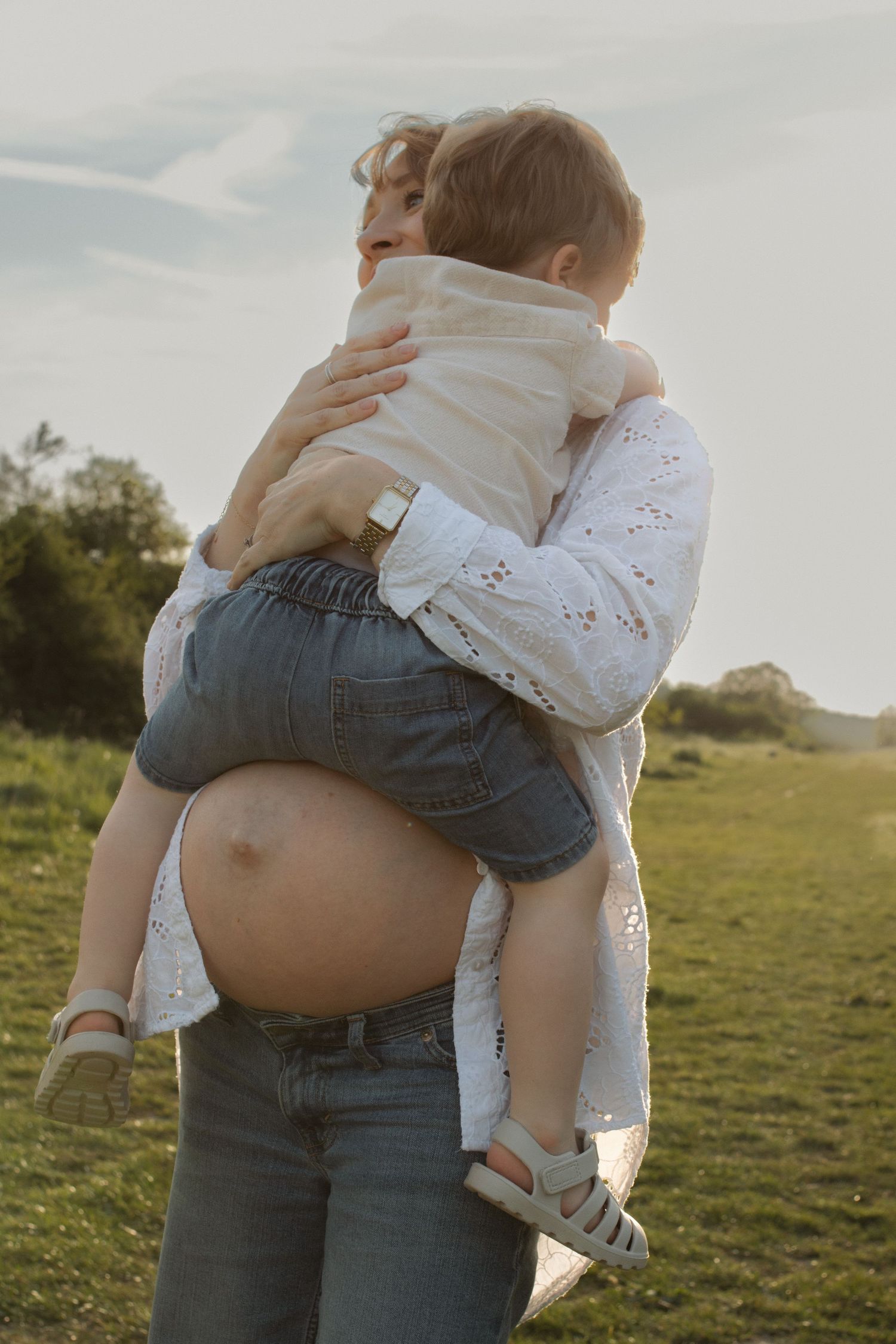 Pregnant woman in white lace top and jeans holds a toddler in an open green field during golden hour.