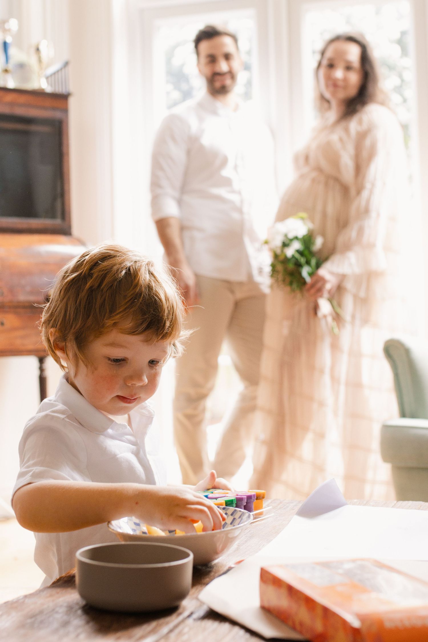 Child coloring at table while couple stands in background by bright window.