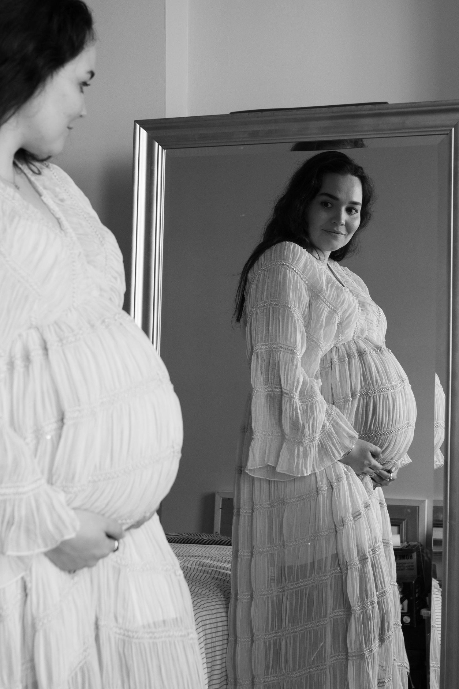 Pregnant woman in white lace dress looking at her reflection in a mirror.