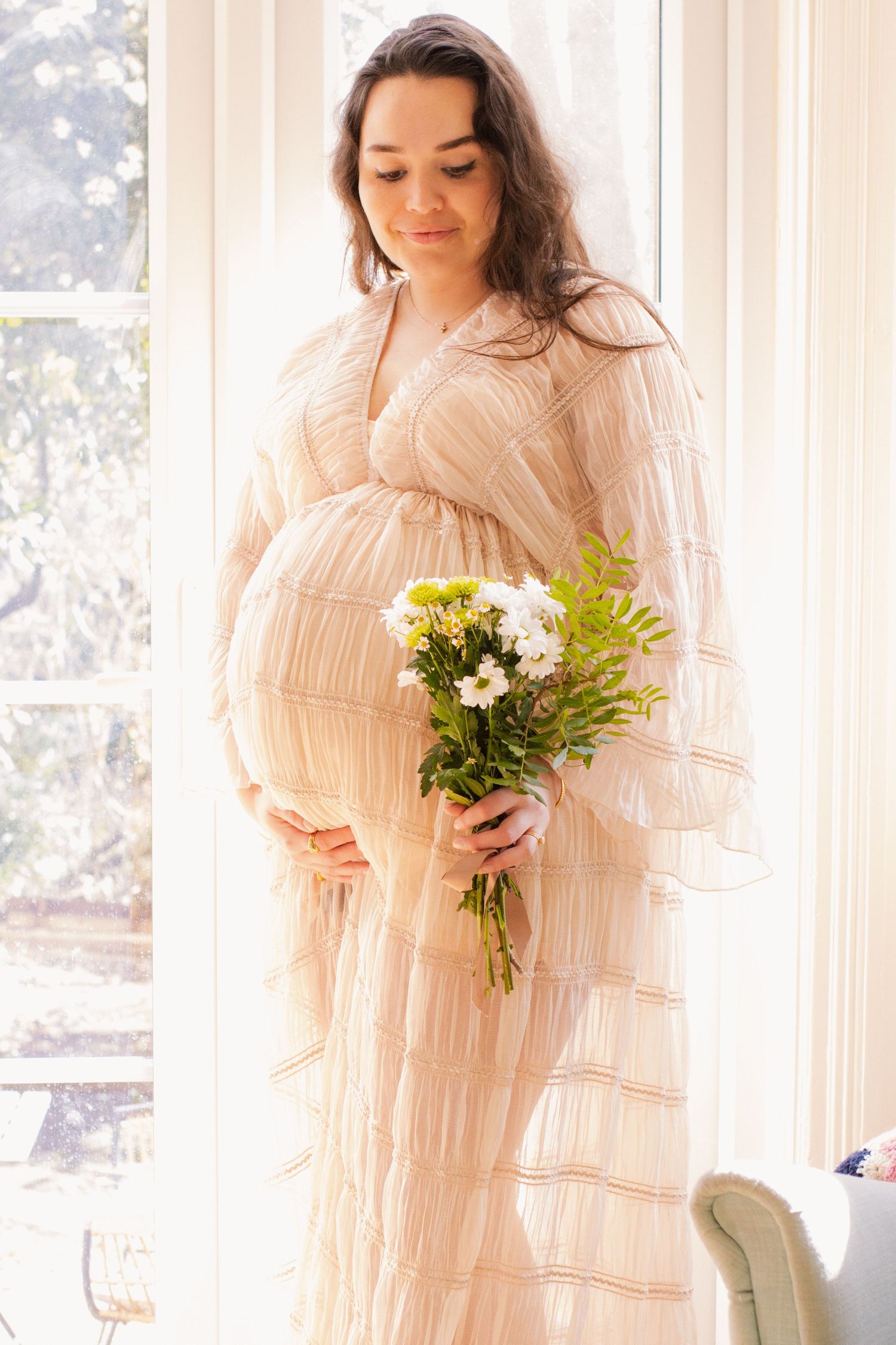 Expectant mother in flowing white dress holds bouquet of flowers by sunlit window.