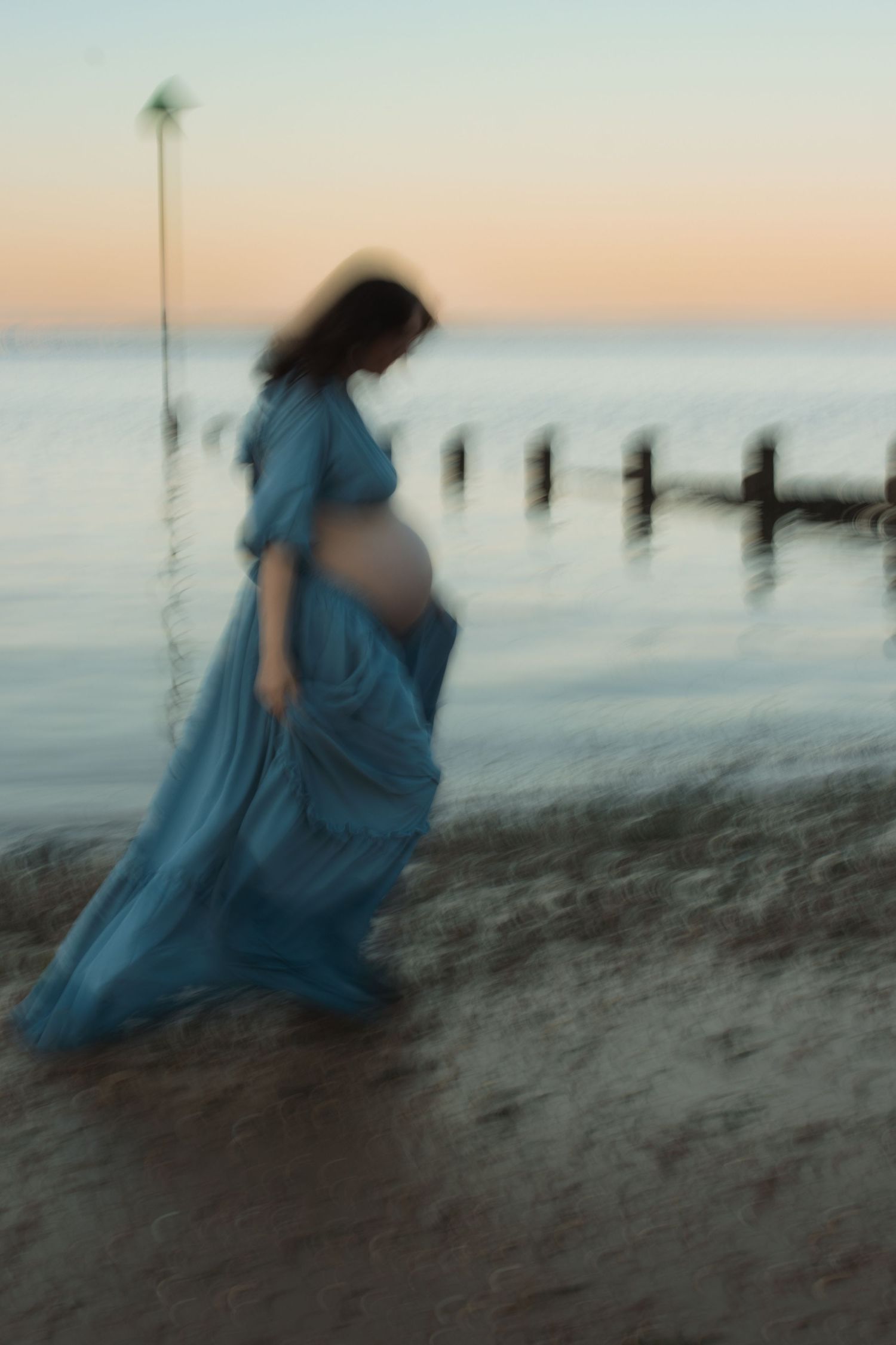 Blurred pregnant woman in flowing blue dress walking on beach at sunset near old wooden pier posts.